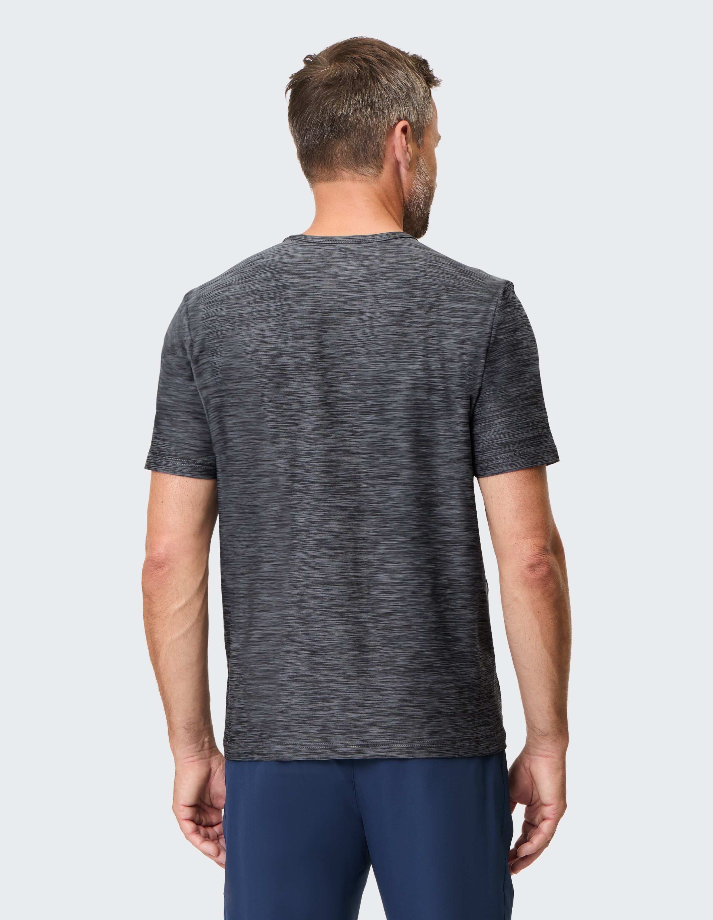 Rückansicht eines Mannes in grauem T-Shirt und blauer Hose, stehend vor hellem Hintergrund, mit kurzem Haar und Bart.