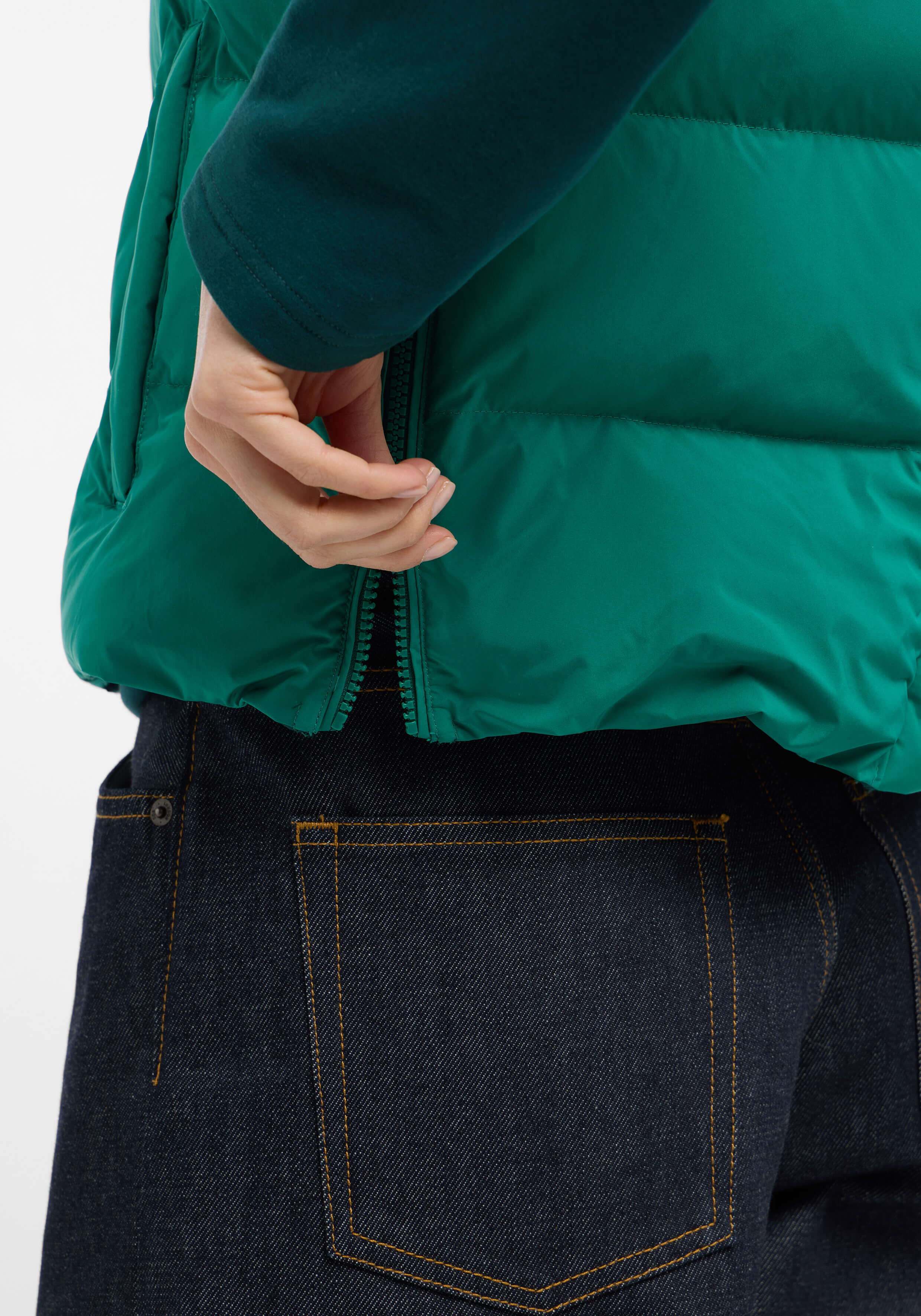 Nahaufnahme einer Hand, die den Reißverschluss einer grünen Daunenweste über einer dunklen Jeans zieht.