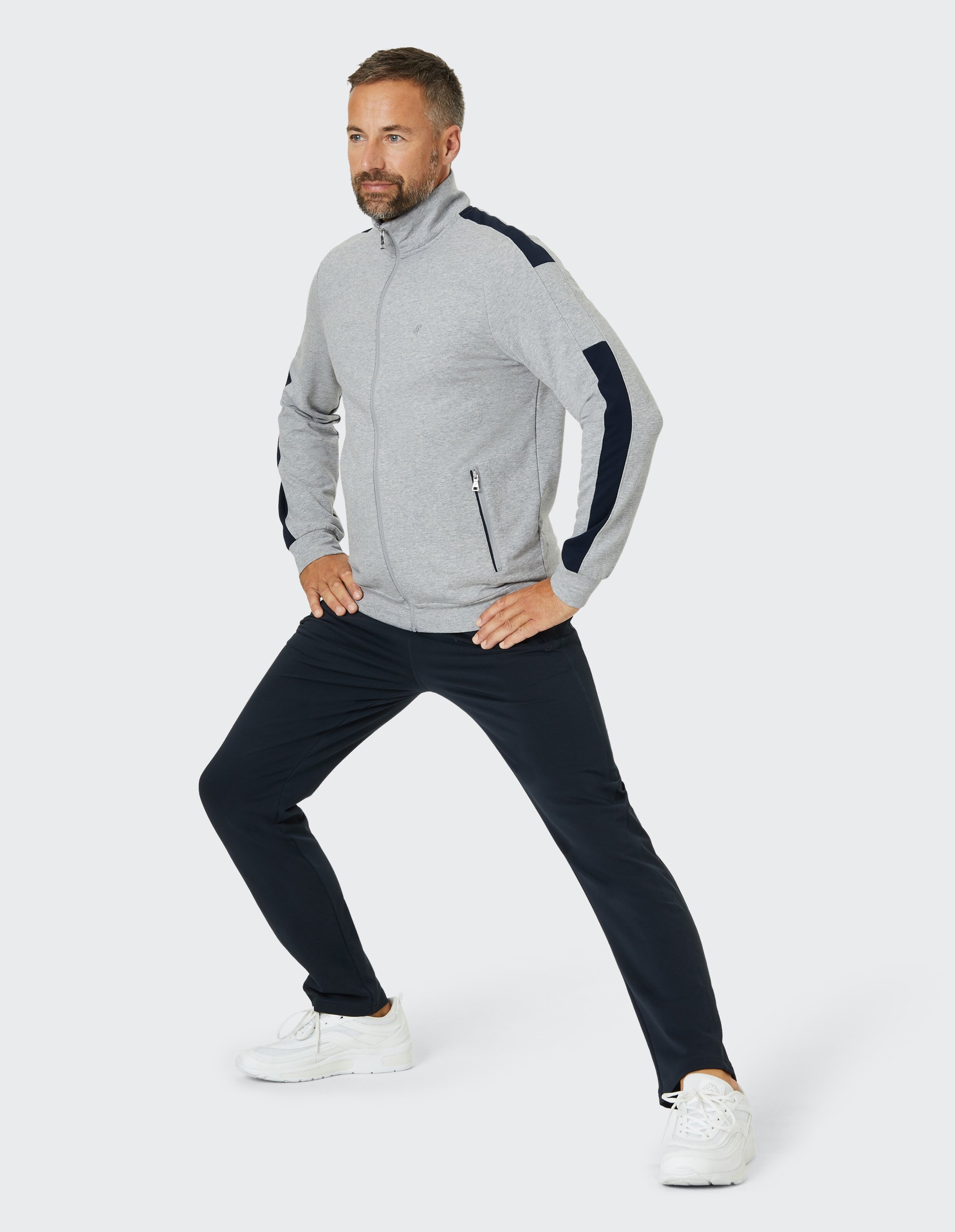 Ein Mann in grauer Jacke und blauen Hosen macht einen Ausfallschritt, Hände auf den Hüften, auf hellem Hintergrund.