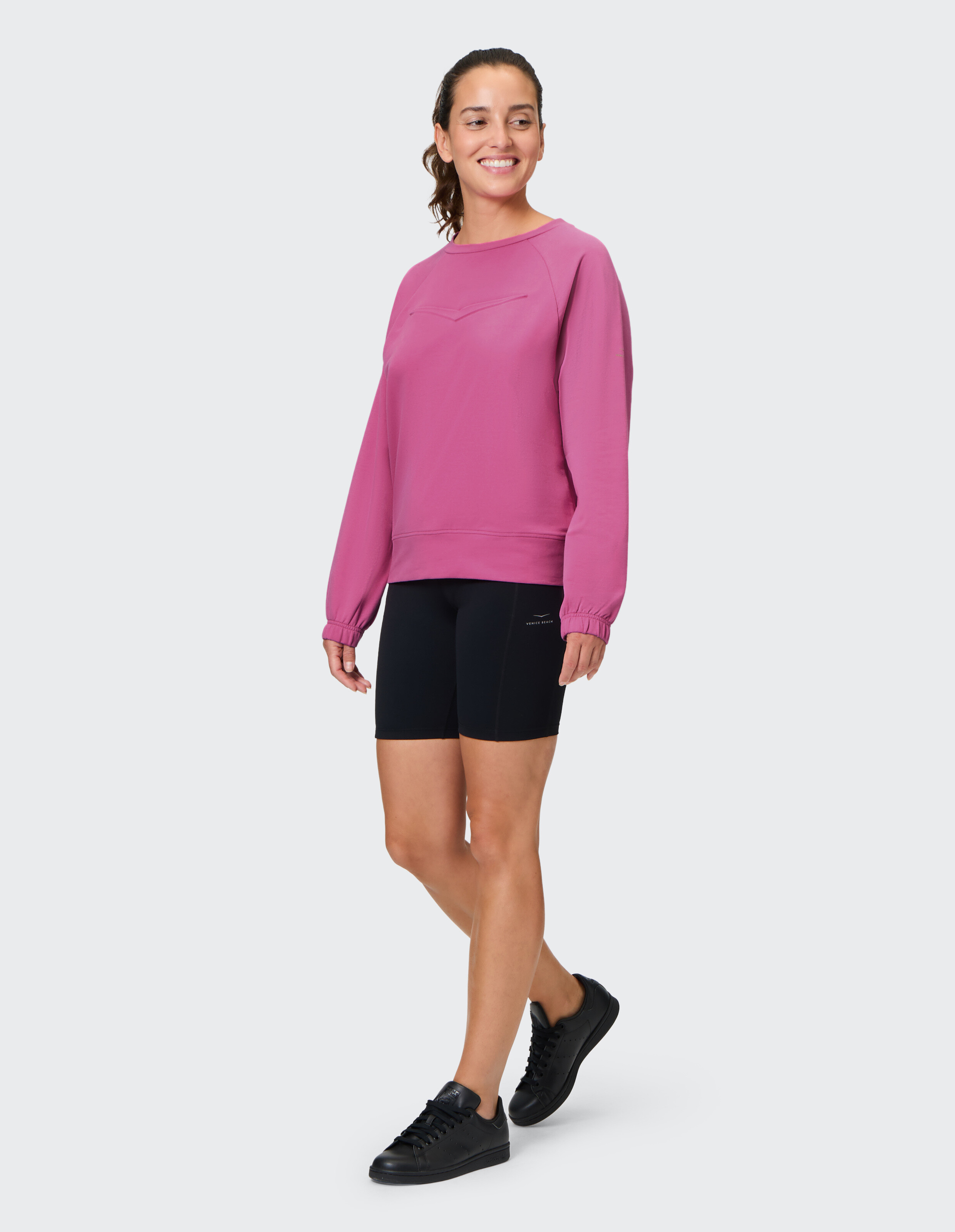 Frau trägt ein rosafarbenes Venice Beach Eartha-Sweatshirt und schwarze Bikershorts.