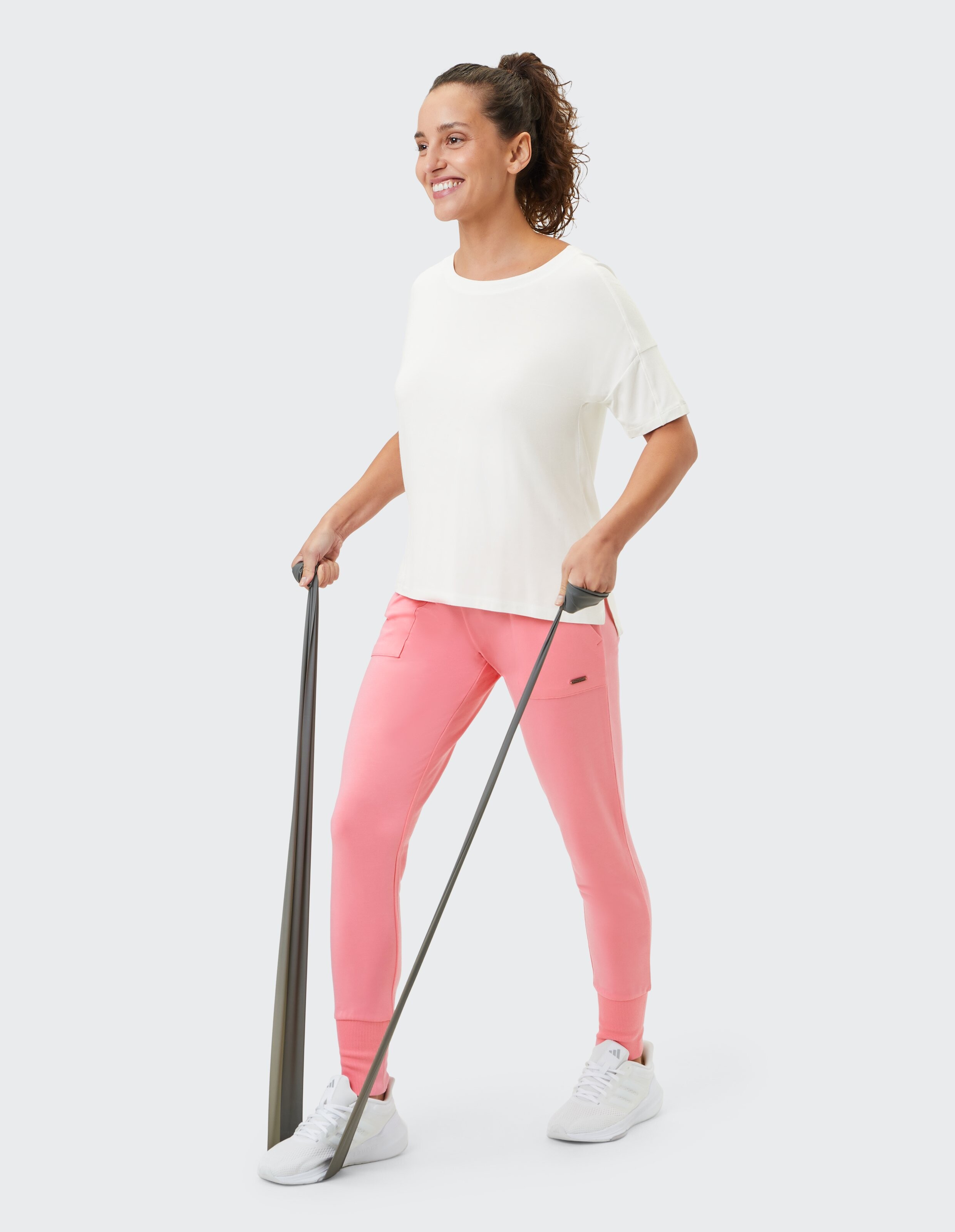 Frau in weißem Top und rosafarbener Jogginghose nutzt Widerstandsbänder für lässige Fitness.