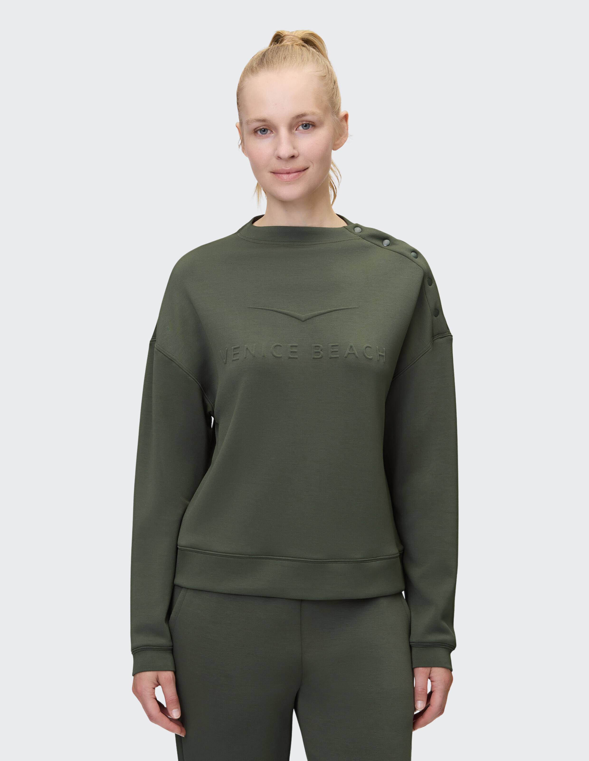 Frau trägt ein gemütliches olivgrünes Sweatshirt mit Knopfdetails und Logo.