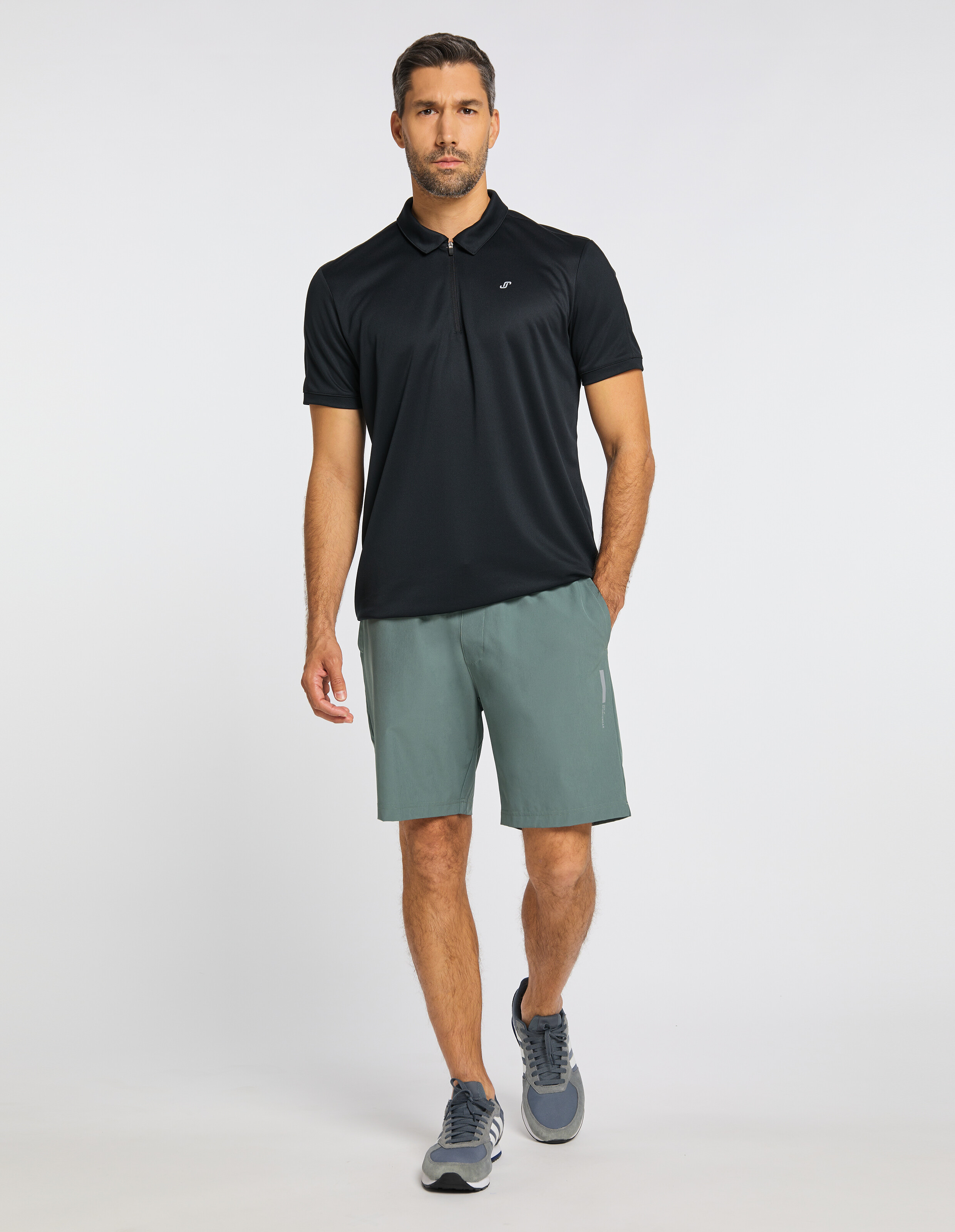 Mann in schwarzem Polohemd und grauen Shorts, der vor hellem Hintergrund steht. Er trägt dunkle Sneaker und schaut freundlich.