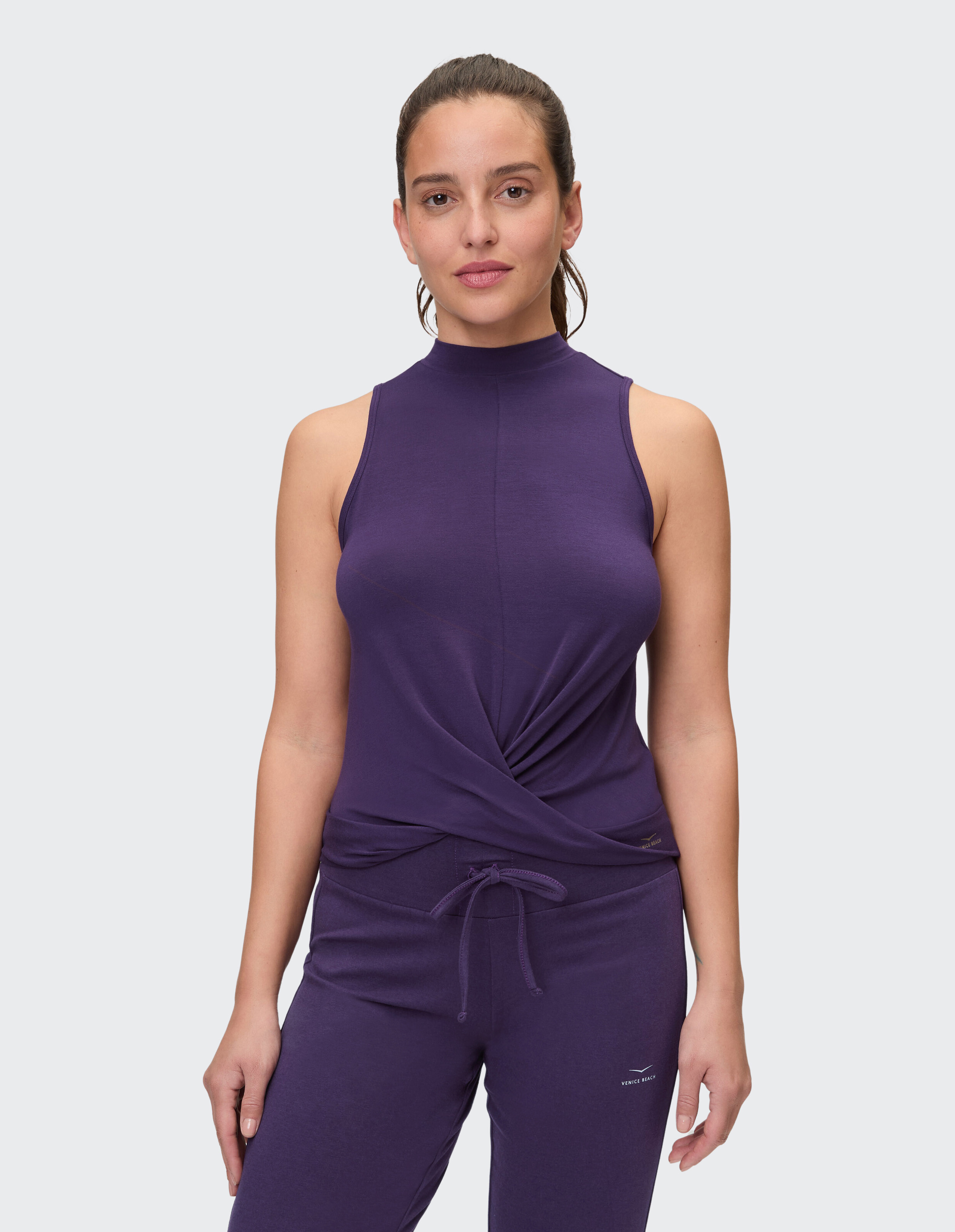 Frau trägt ein lilafarbenes Venice Beach Aria-Tanktop mit Stehkragen und Twist-Detail vorne.
