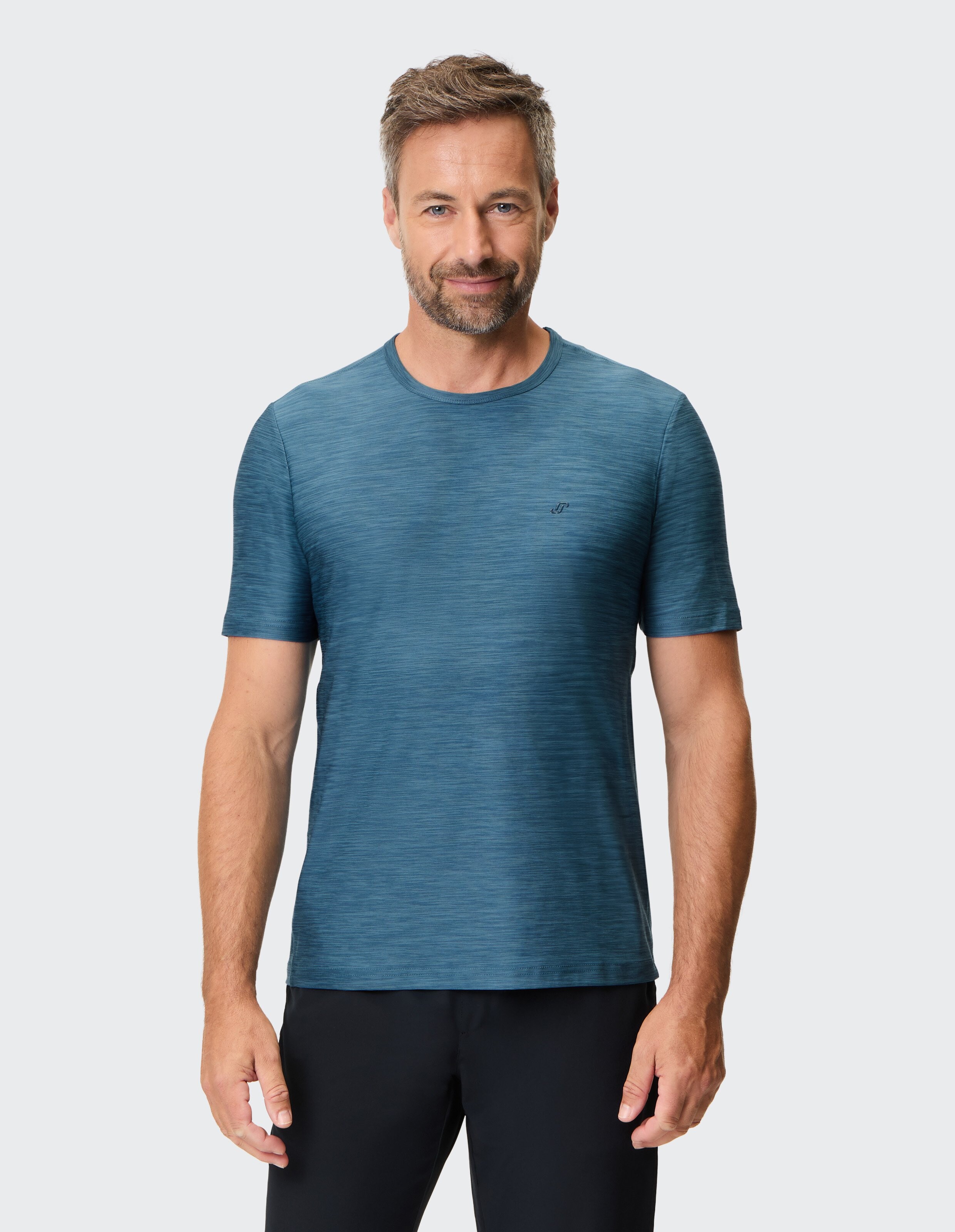 Ein lächelnder Mann mit kurzem Haar trägt ein blaues T-Shirt und schwarze Hose vor einem schlichten, weißen Hintergrund.