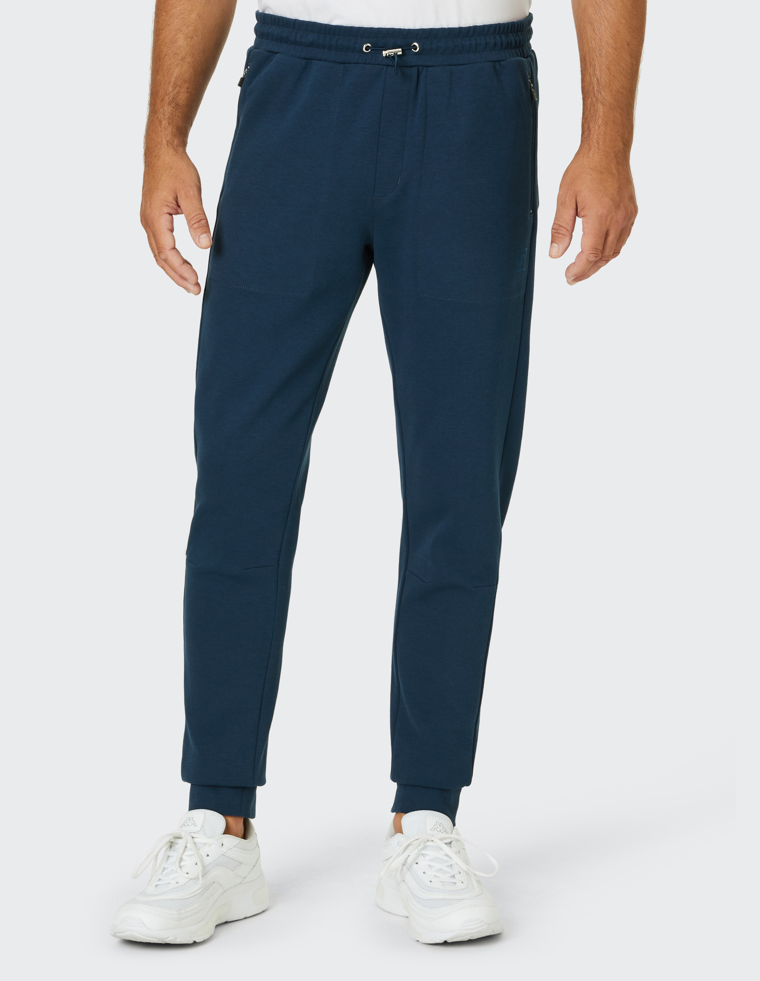 Ein Mann trägt eine dunkelblaue Jogginghose mit Kordelzug und weiße Turnschuhe auf hellem Hintergrund, Oberkörper nicht sichtbar.