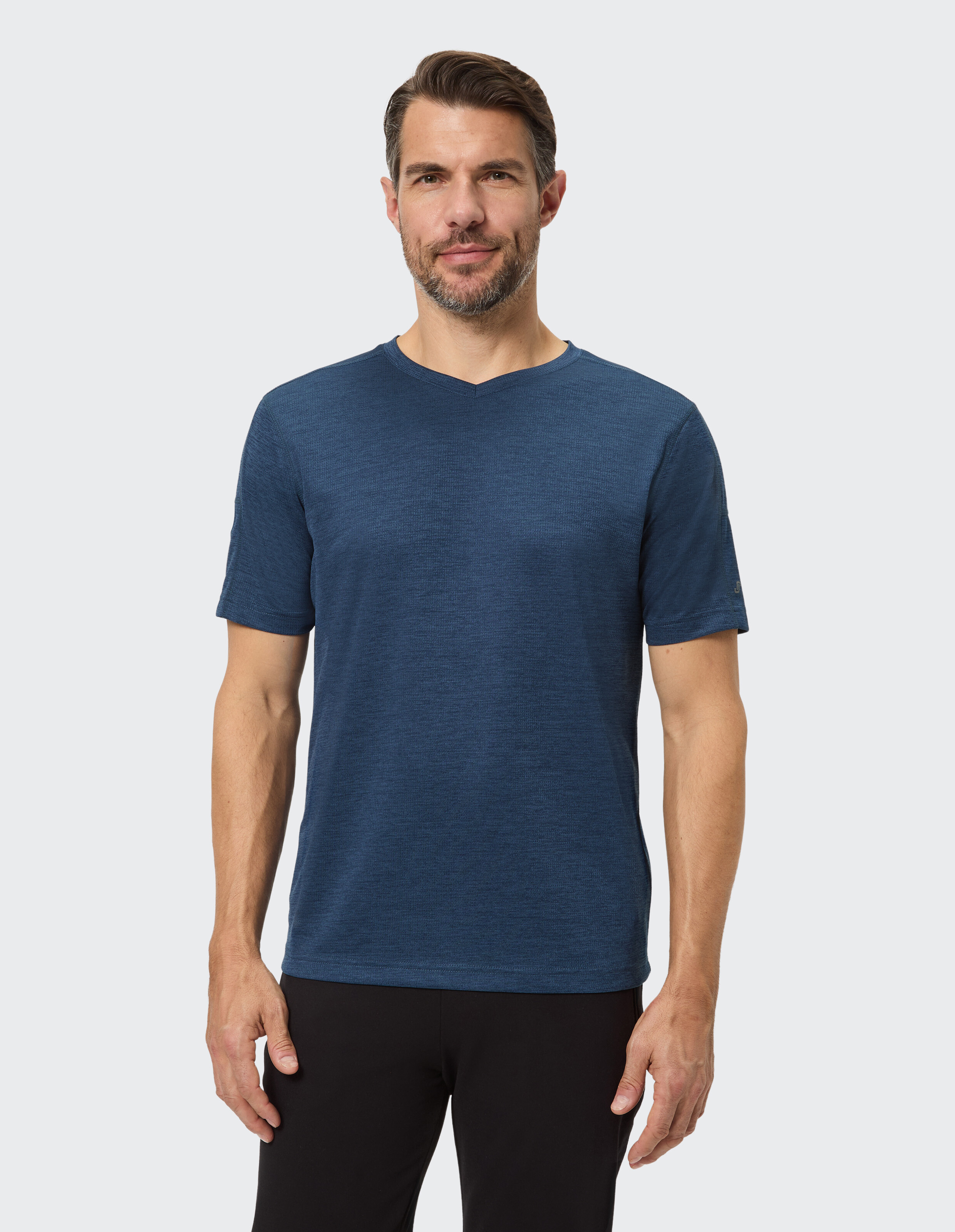 Ein Mann trägt ein blaues T-Shirt und schwarze Hose, steht vor einem hellgrauen Hintergrund und lächelt leicht in die Kamera.