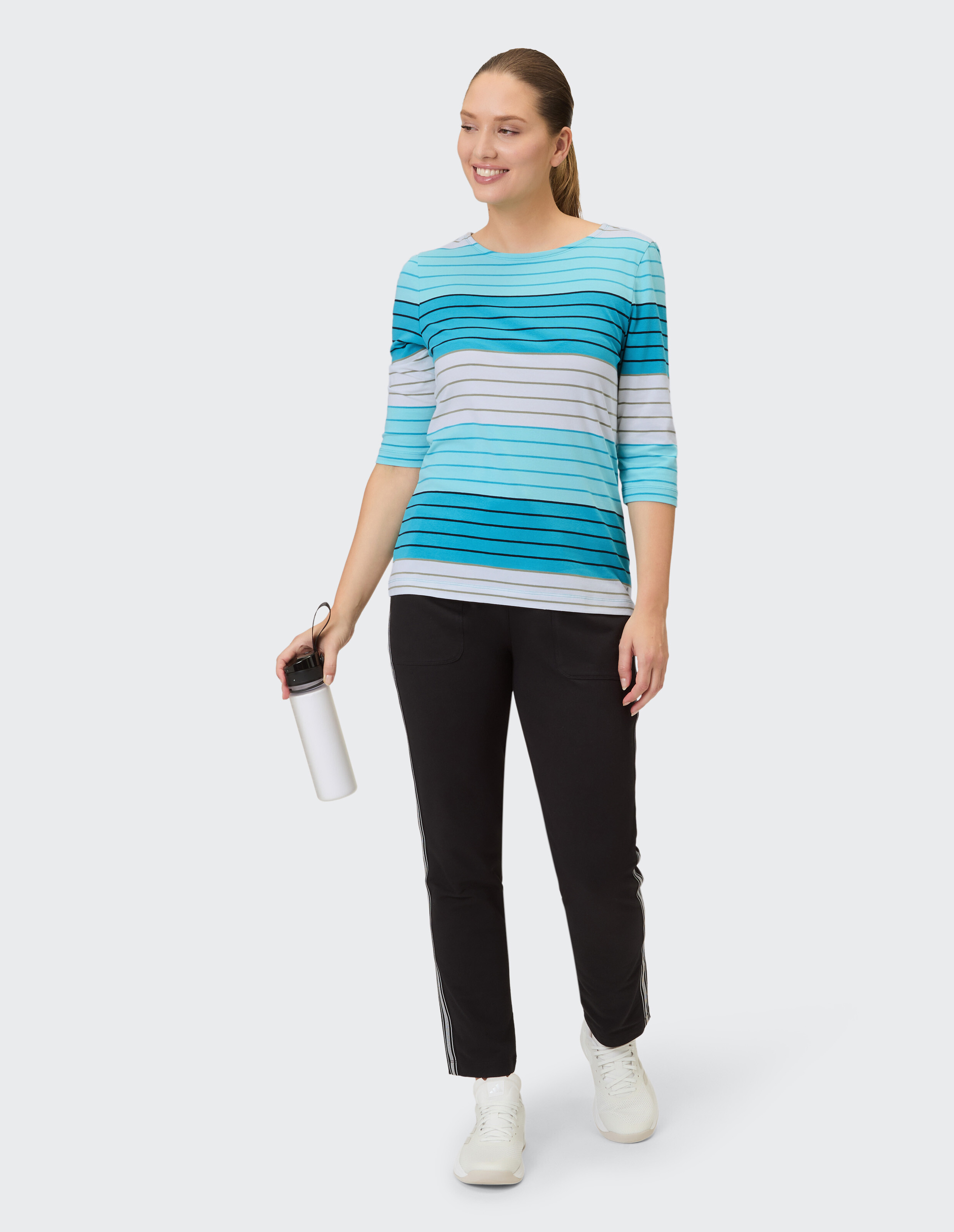 Frau in gestreiftem blauen Top und schwarzer Hose, eine Wasserflasche in der Hand, lässiger Stil.
