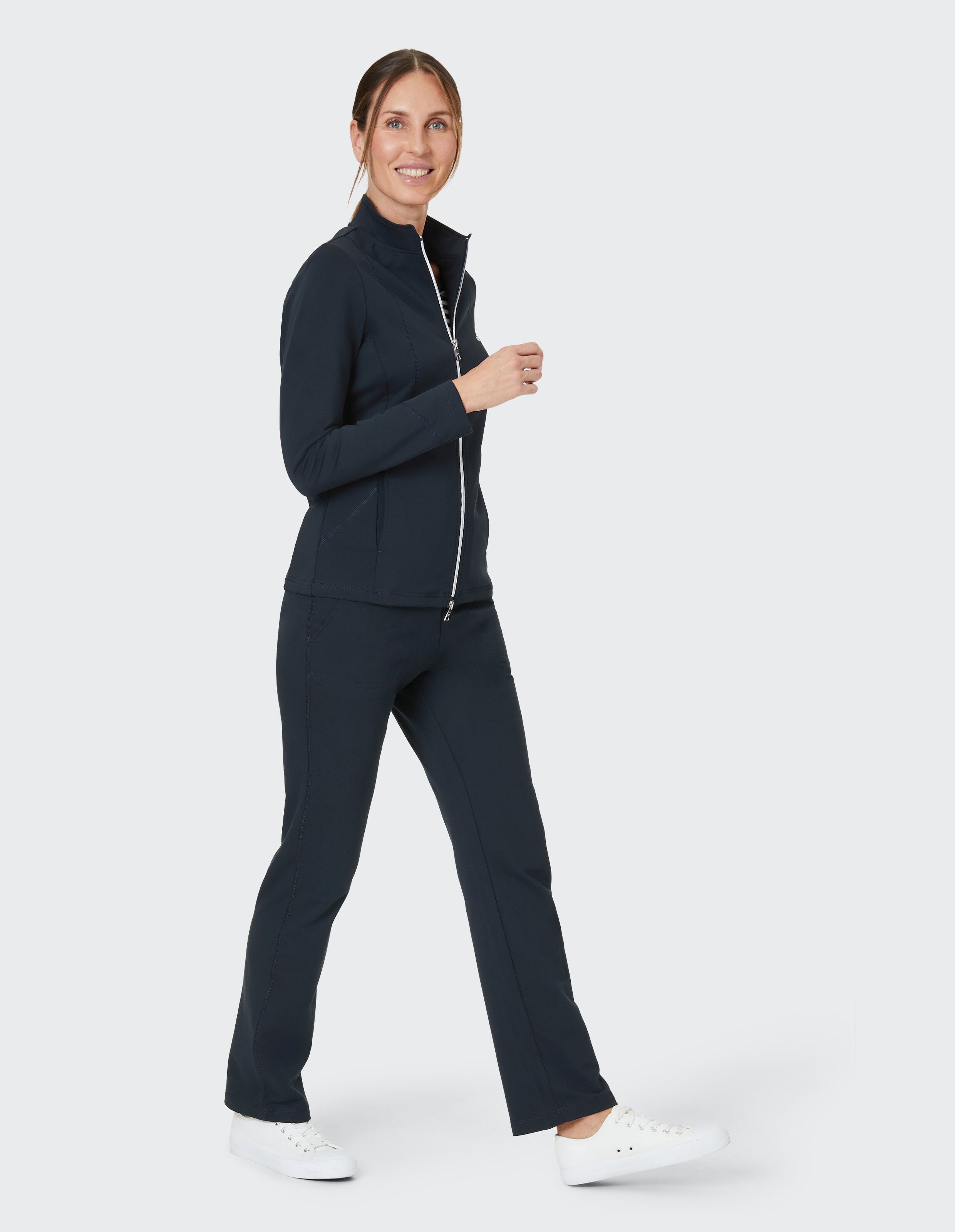Frau in dunkelblauem Trainingsanzug und weißen Sneakers geht lächelnd vor einem weißen Hintergrund.