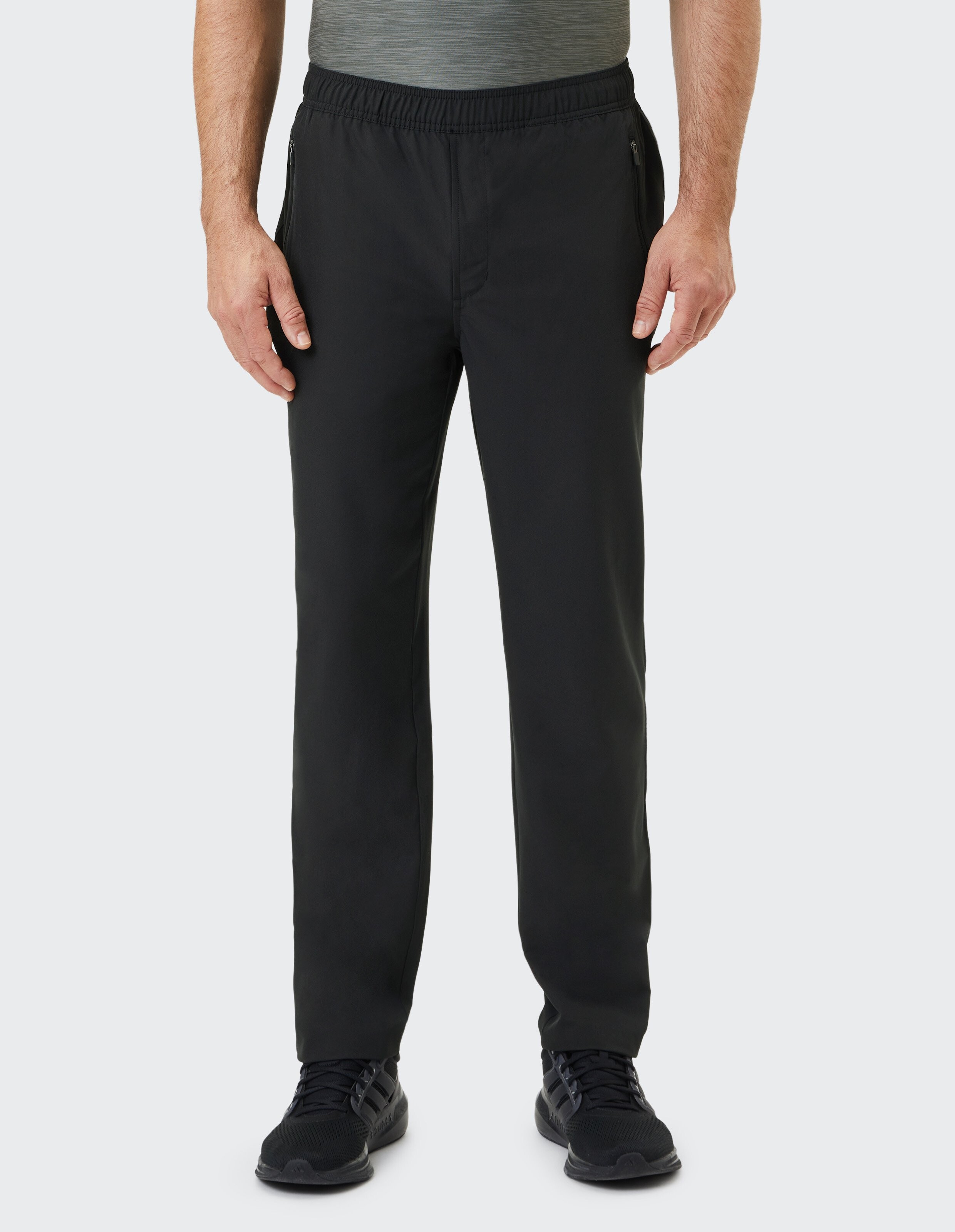 Mann in schwarzen Jogginghosen und weißen Sneakers, stehend, mit einer Hand in der Hosentasche; weißer Hintergrund.