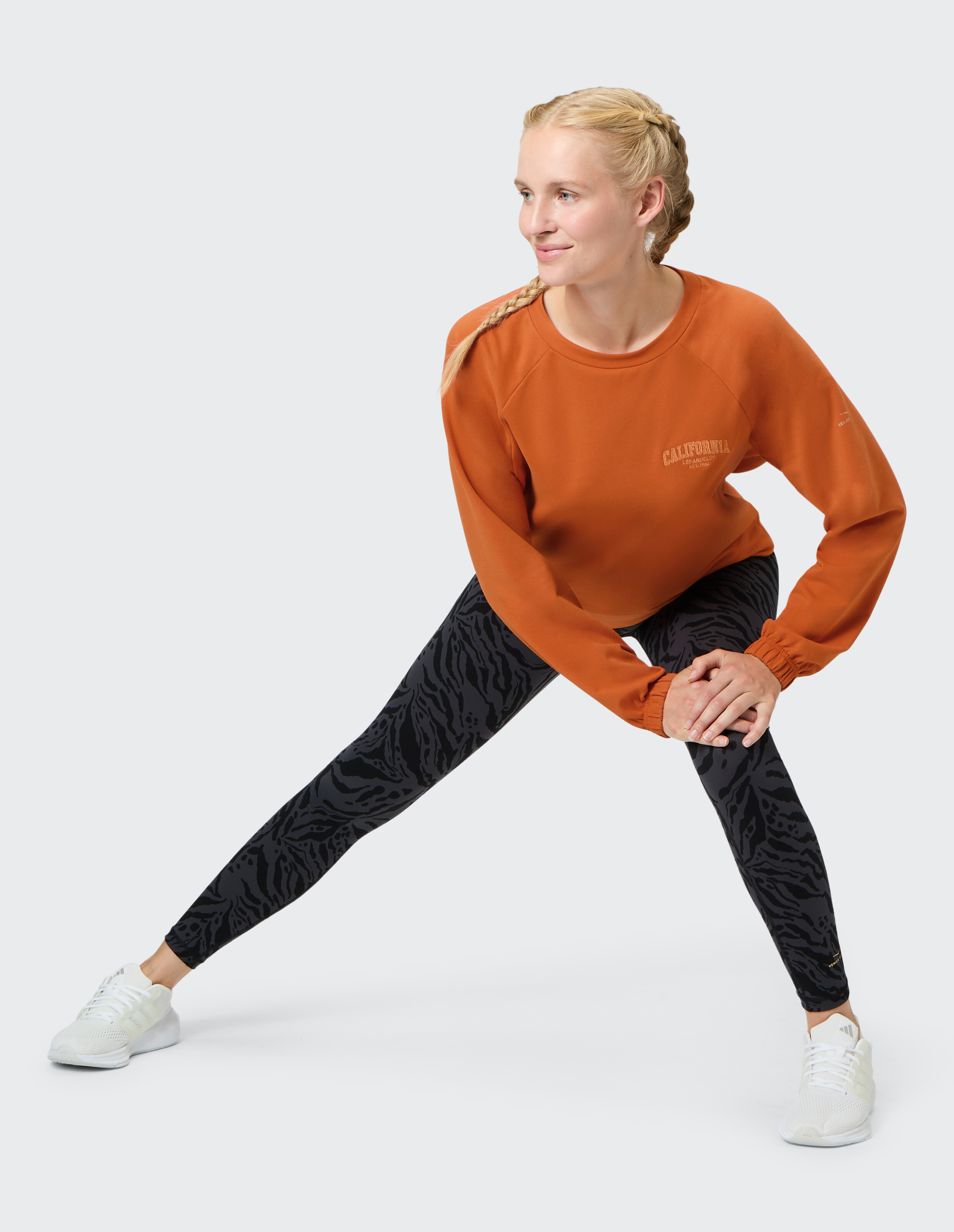 Frau beim Stretching in einem orangefarbenen Sweatshirt und schwarz gemusterten Leggings, ideal für Freizeitkleidung.