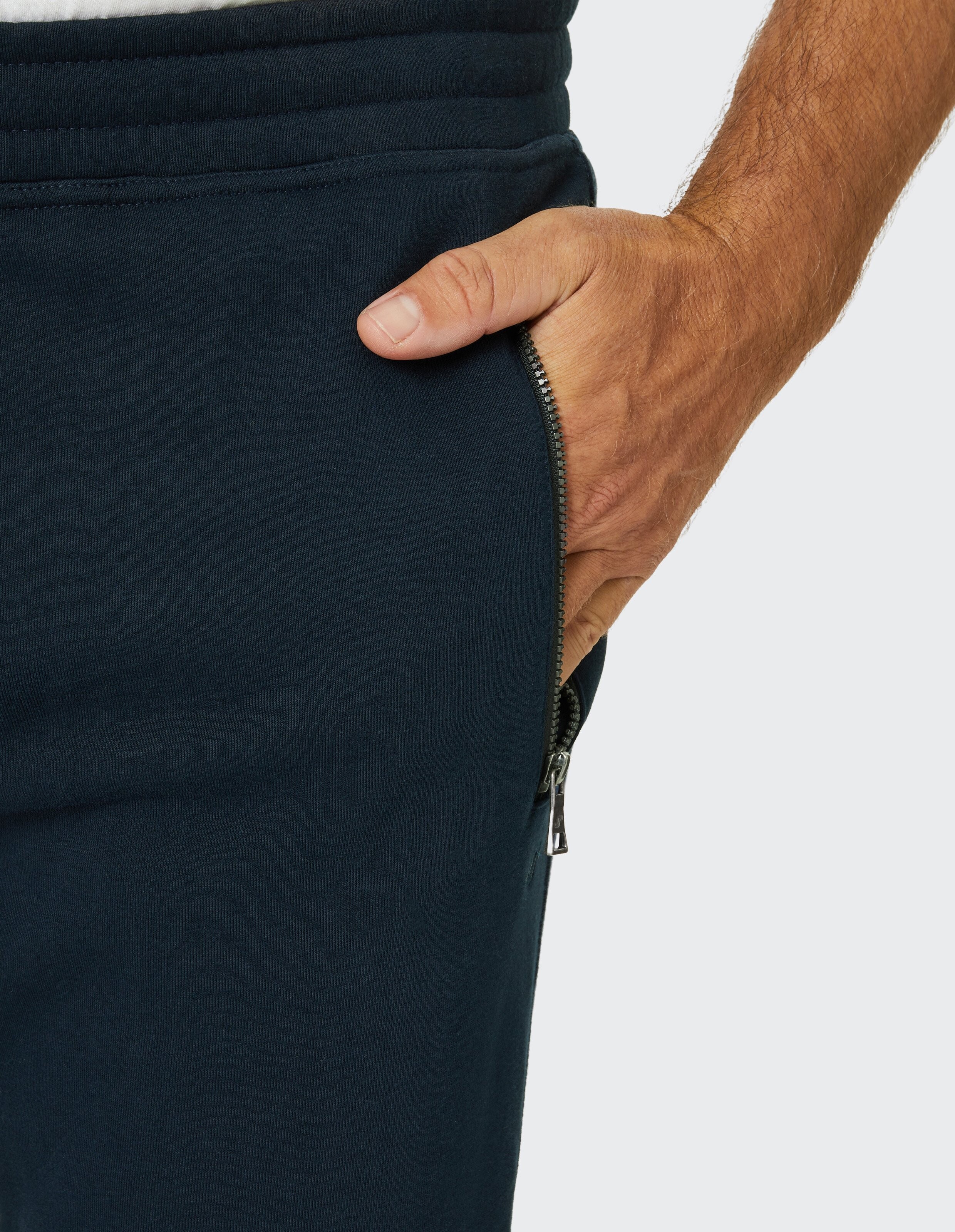 Eine Hand steckt in die Seitentasche einer dunkelblauen Jogginghose mit Reißverschluss, die an einer Person getragen wird.