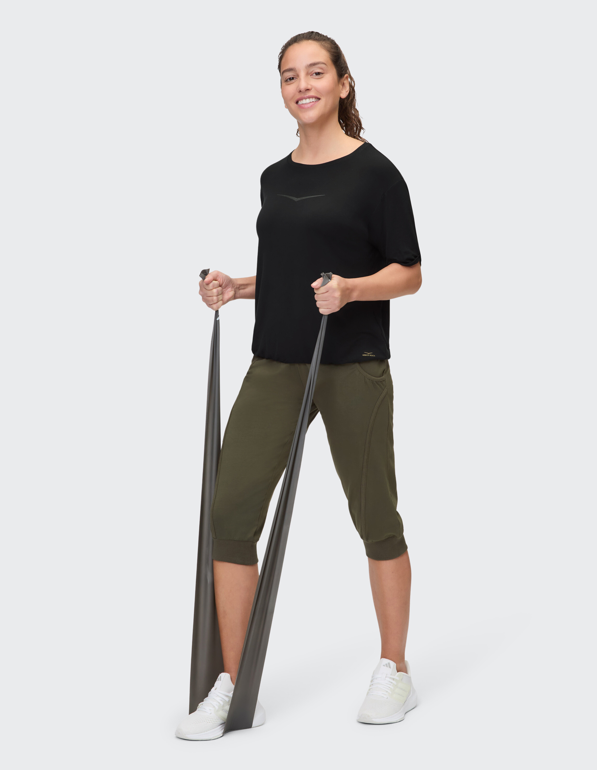 Frau in schwarzem Top und olivgrüner Caprihose, hält Trainingsbänder in der Hand.