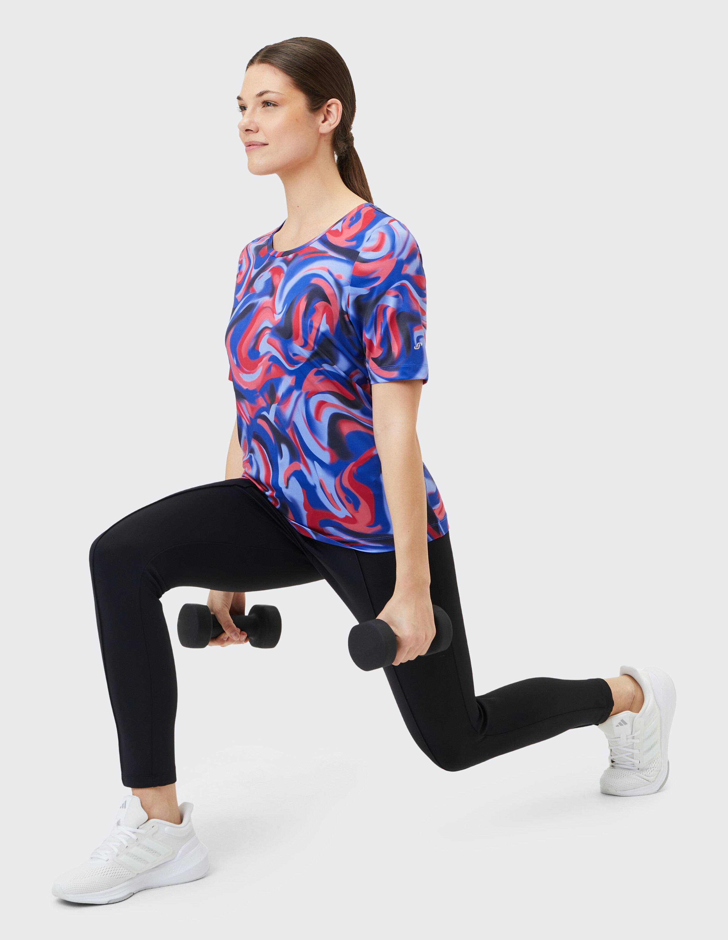 Frau in buntem Sporttop und schwarzen Leggings führt einen Ausfallschritt mit Kurzhanteln aus.