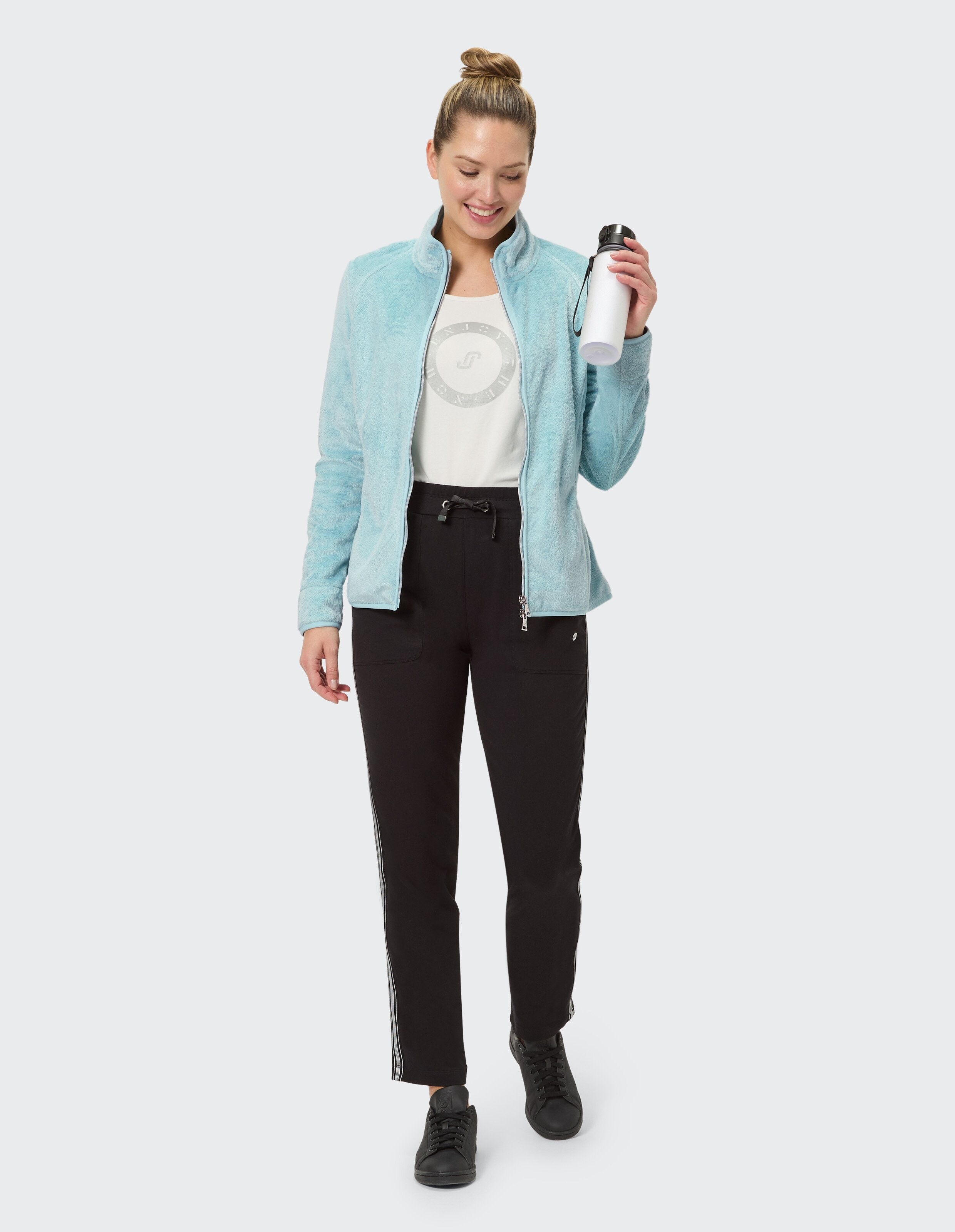 Frau in hellblauer Fleecejacke und schwarzer Jogginghose, hält eine Wasserflasche in der Hand.