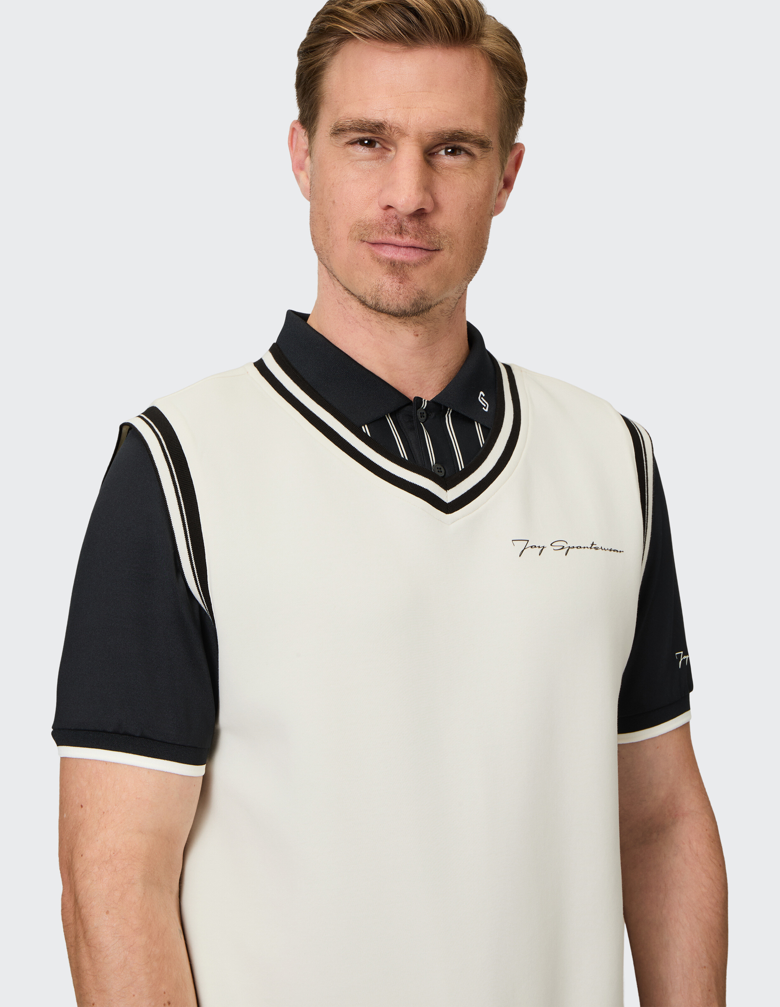 Ein Mann trägt ein cremefarbenes ärmelloses Oberteil mit schwarzem Kragen und Streifen, über einem dunkelblauen Poloshirt.