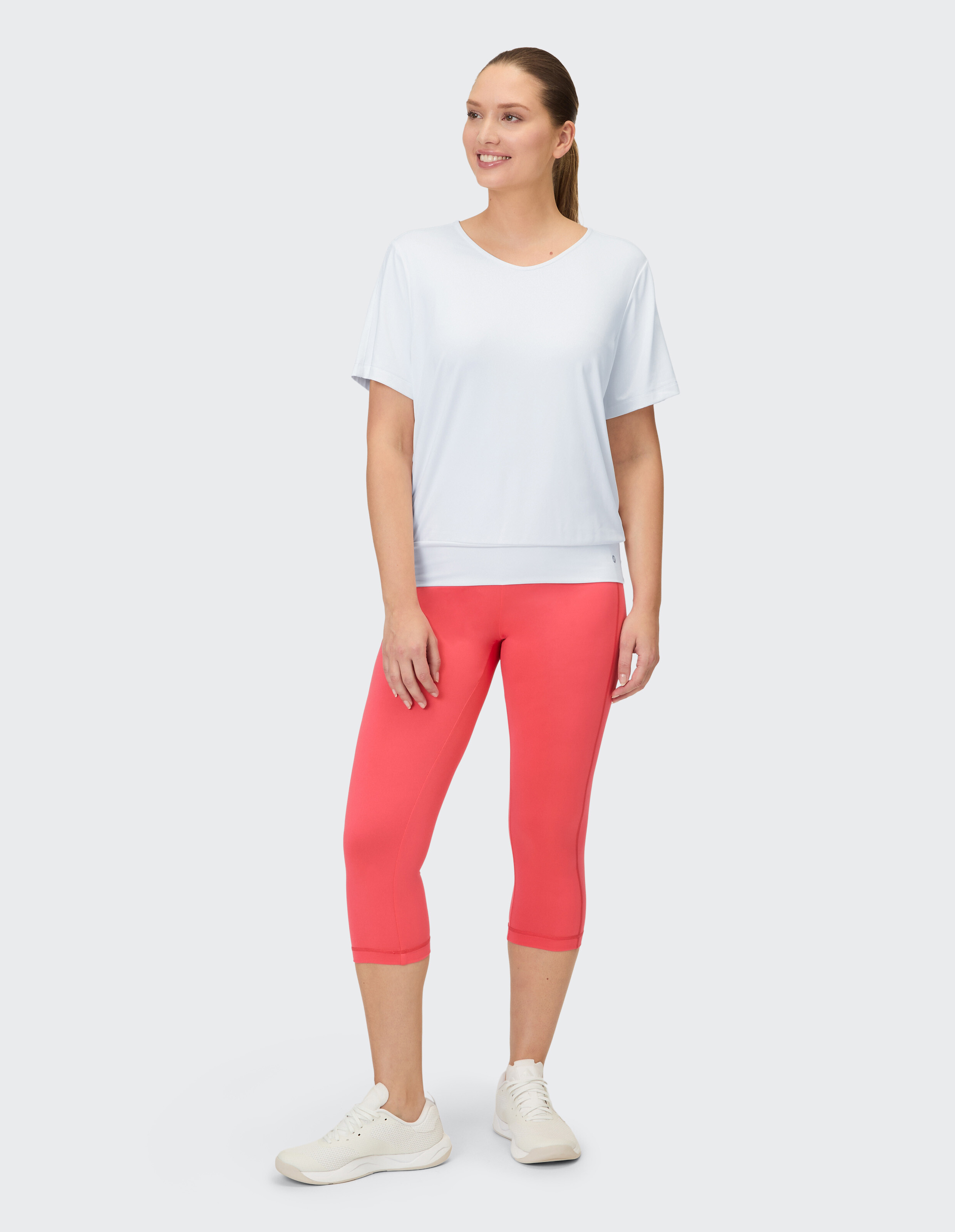 Eine Frau trägt ein weißes Kurzarmshirt und korallenrote 3/4-Leggings.