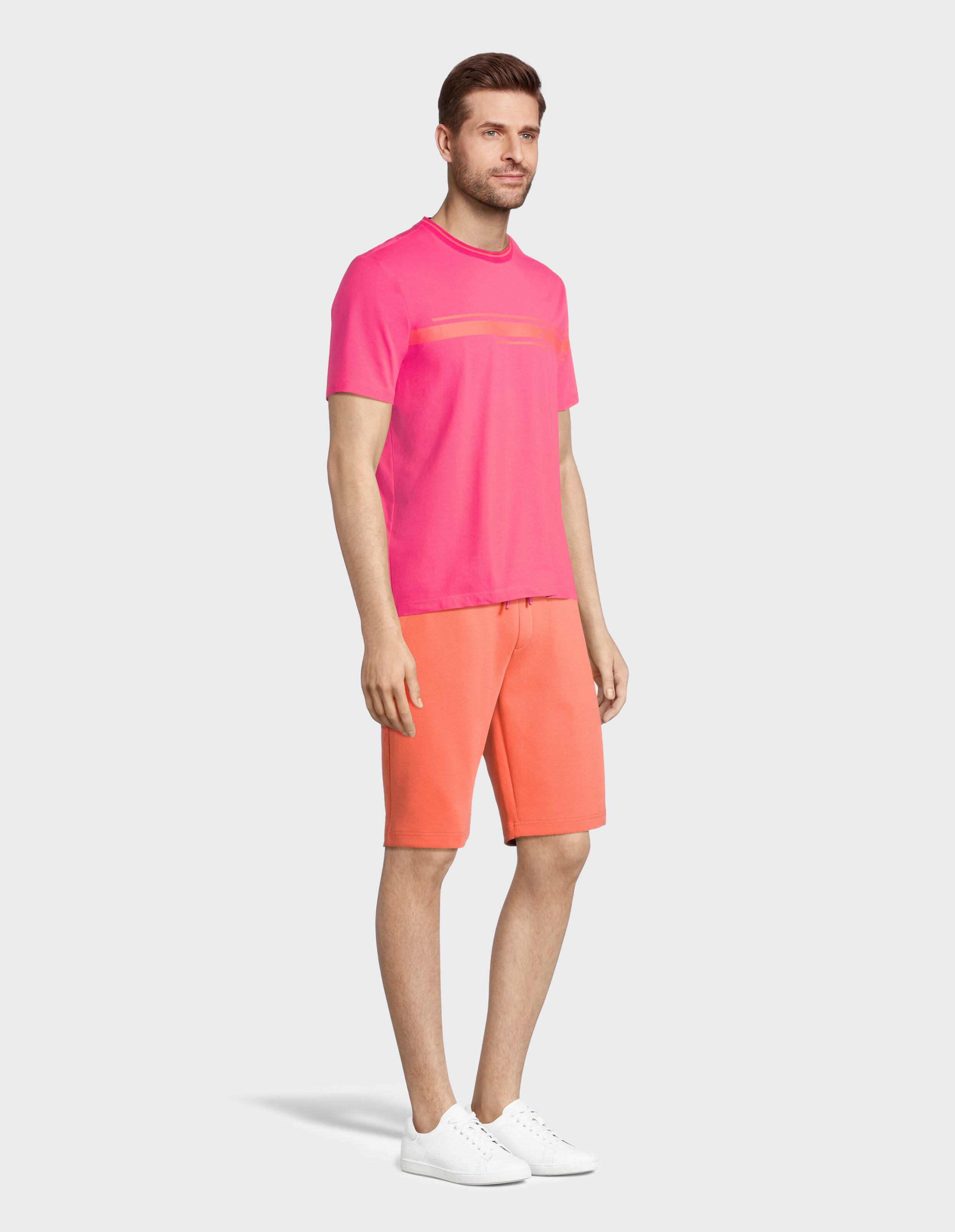 Ein Mann trägt ein pinkes T-Shirt und orange Shorts mit weißen Sneakers vor grauem Hintergrund.