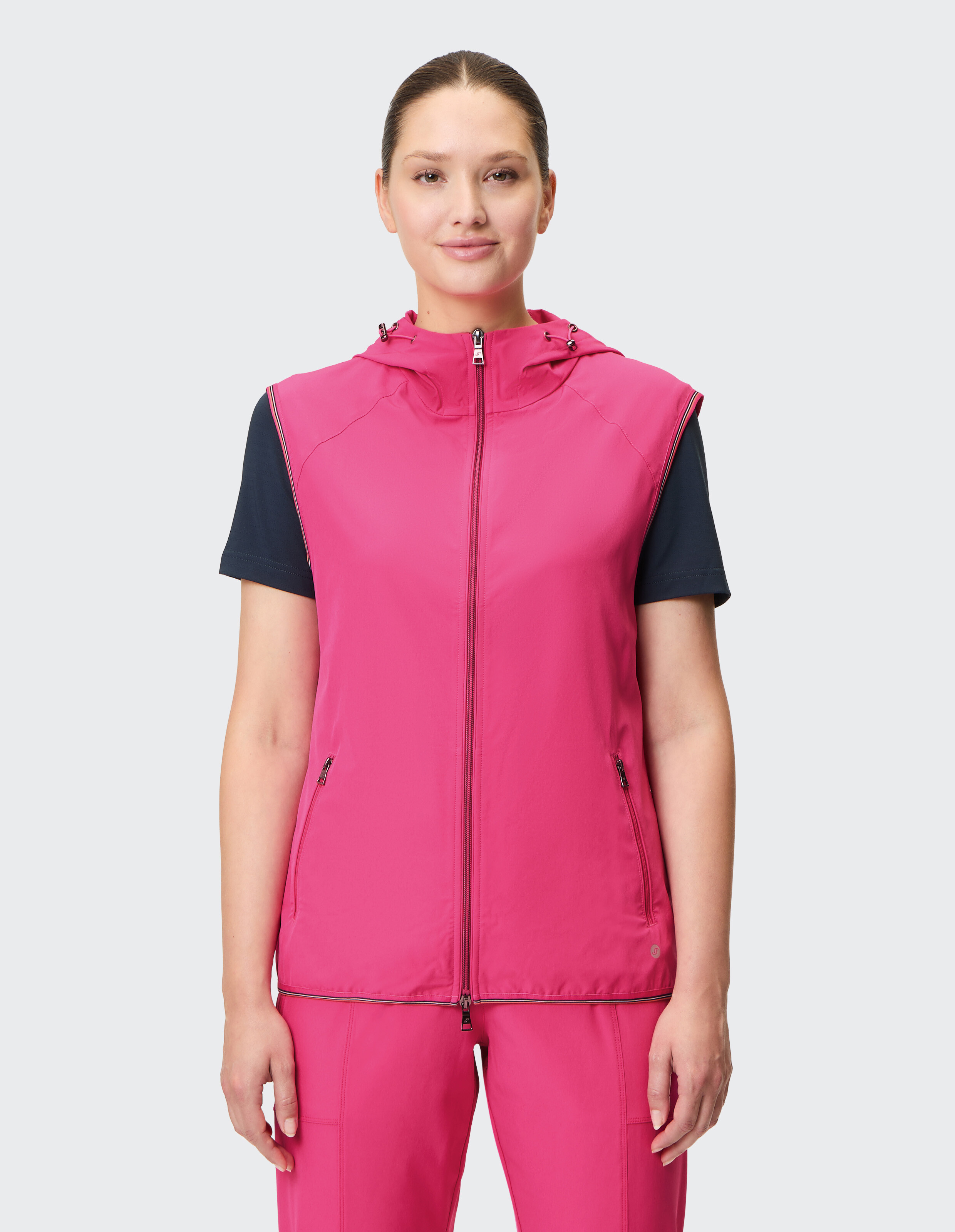 Eine Frau präsentiert eine leuchtend rosa ärmellose Weste mit Kapuze, gepaart mit einem dunkelblauen Hemd, ideal für Freizeitausflüge.
