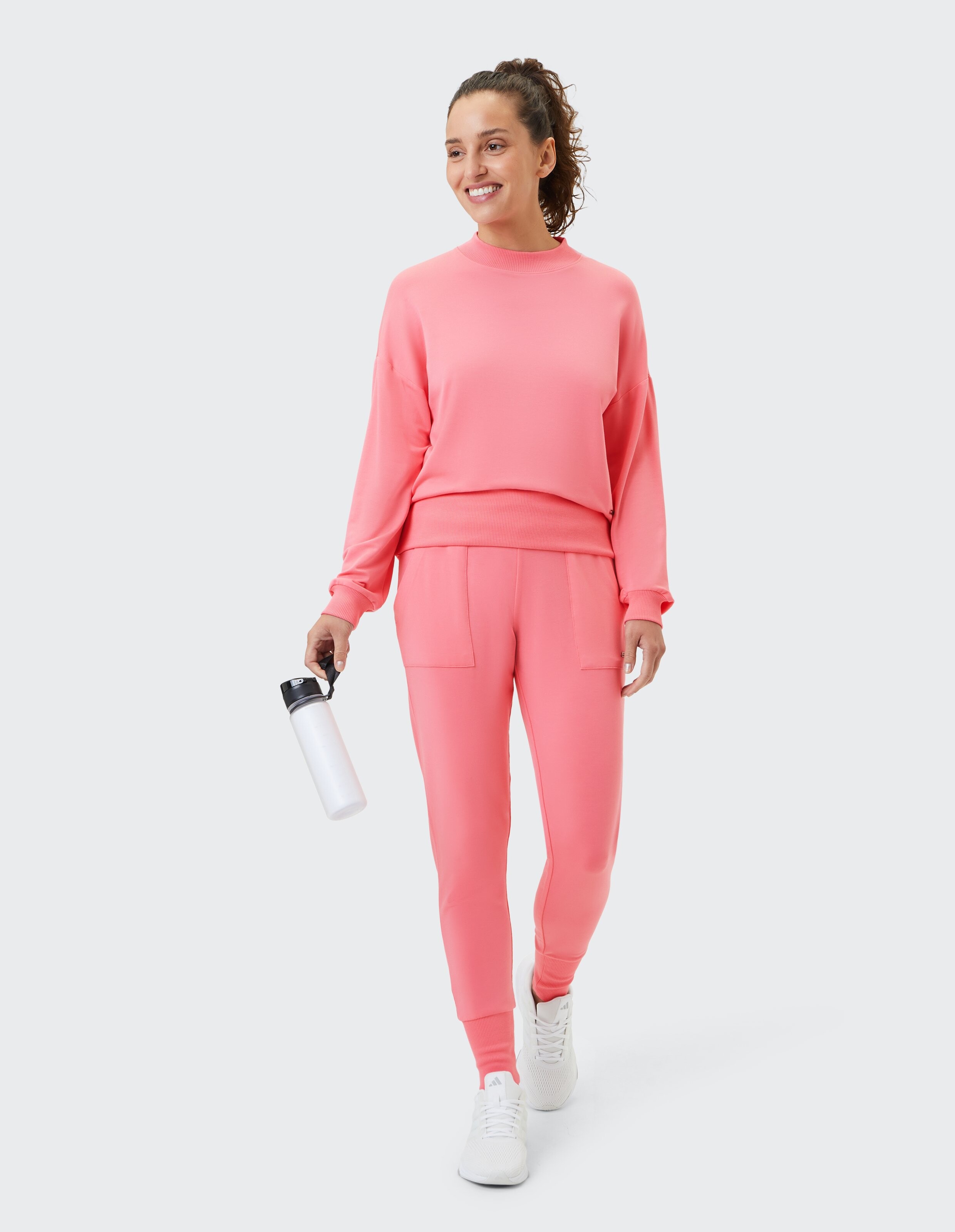 Frau in einem rosa Sweatshirt und passenden Jogginghosen, hält eine Wasserflasche in der Hand.