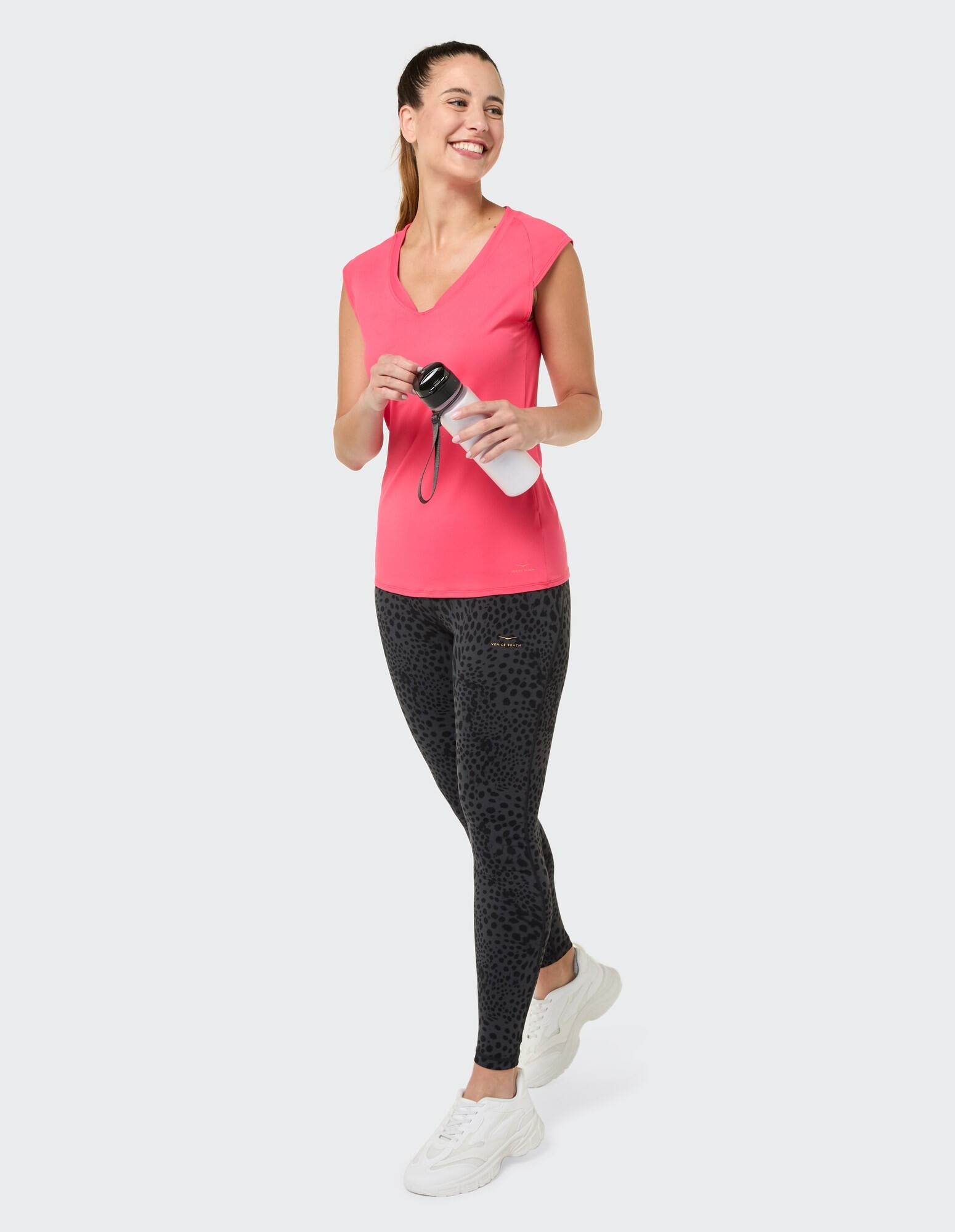 Frau in einem rosafarbenen Top mit V-Ausschnitt und schwarzen Leggings mit Leopardenmuster, hält eine Wasserflasche in der Hand.