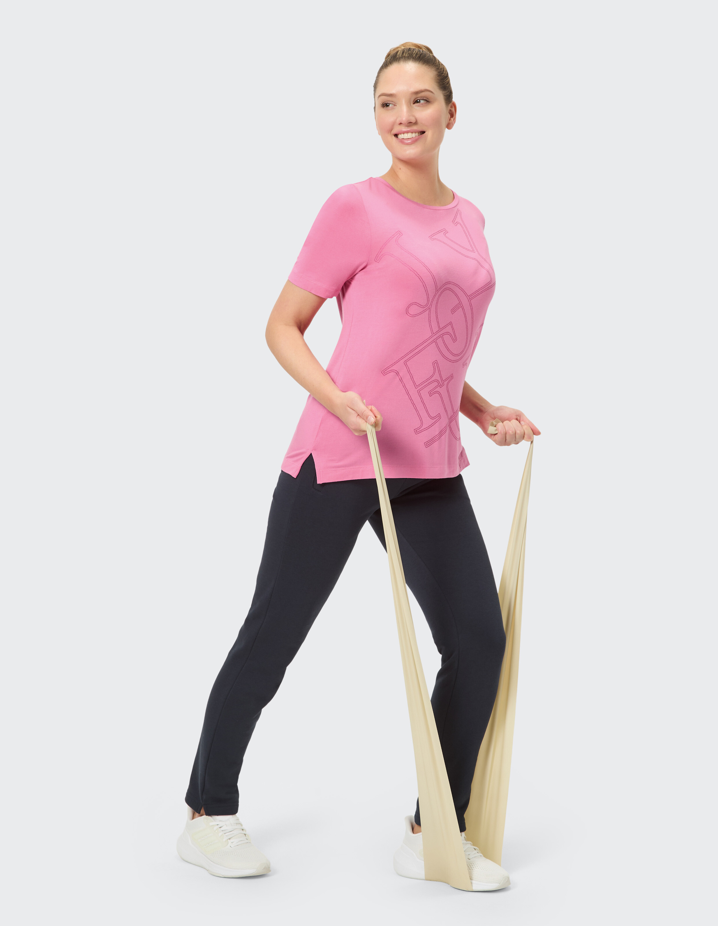 Frau im rosa T-Shirt mit "JOYFUL"-Aufdruck trägt dunkelblaue Hose und weiße Turnschuhe, lächelnd vor neutralem Hintergrund.