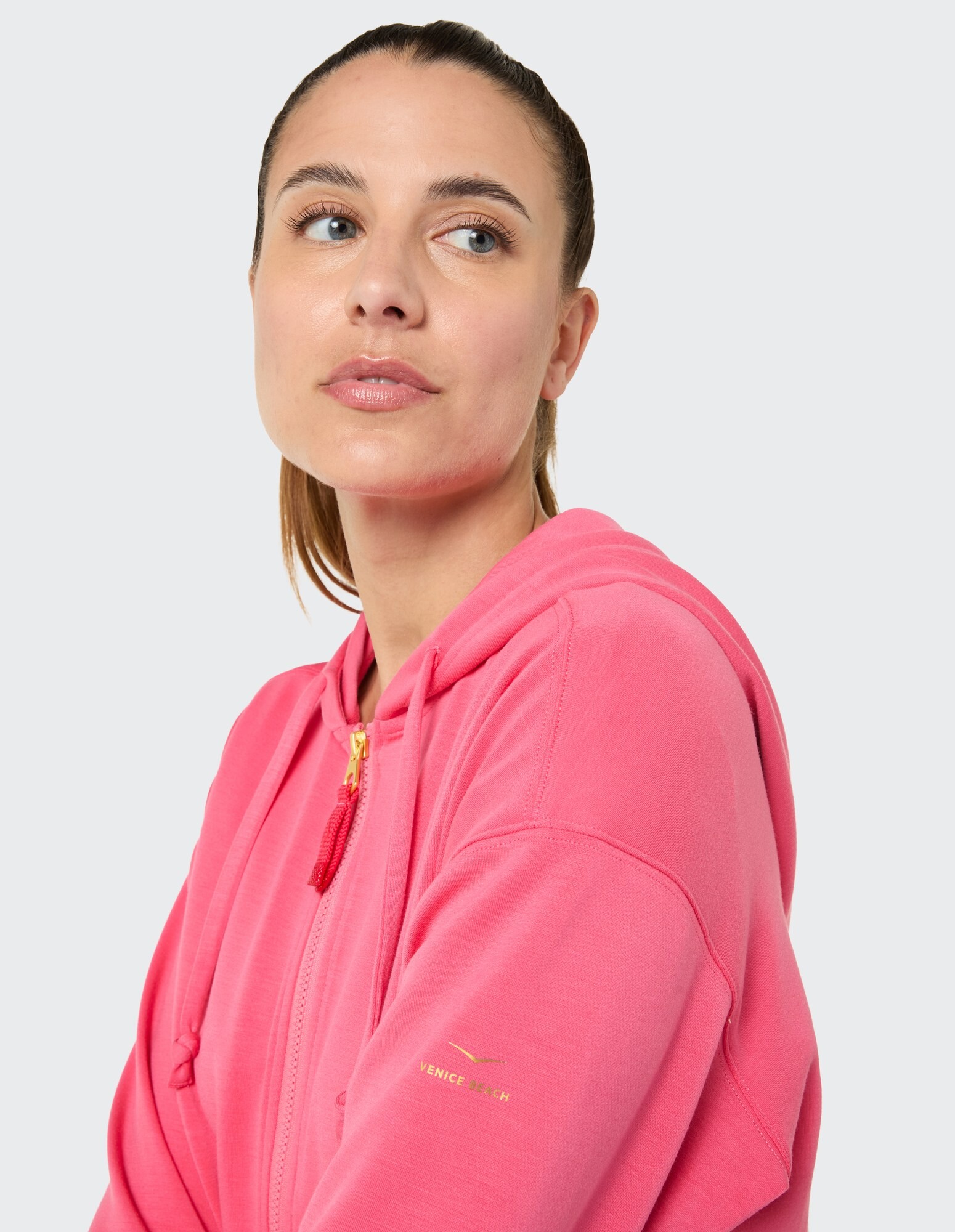 Das Model trägt einen rosa Kapuzenpullover mit Reißverschluss im lässigen Stil und goldenem Logo-Detail.