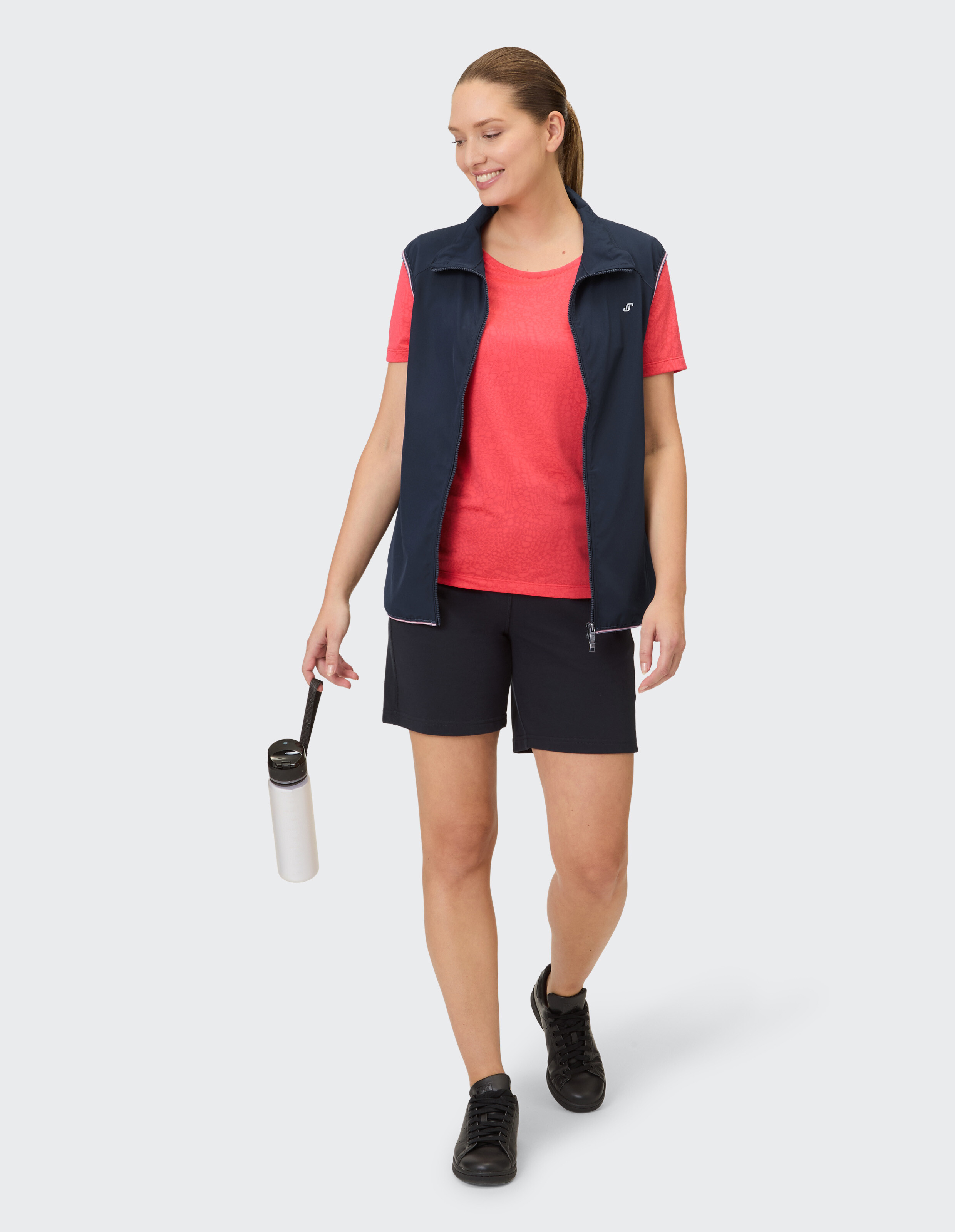 Frau in marineblauer Weste, rotem Hemd und schwarzen Shorts, hält eine Wasserflasche in der Hand.