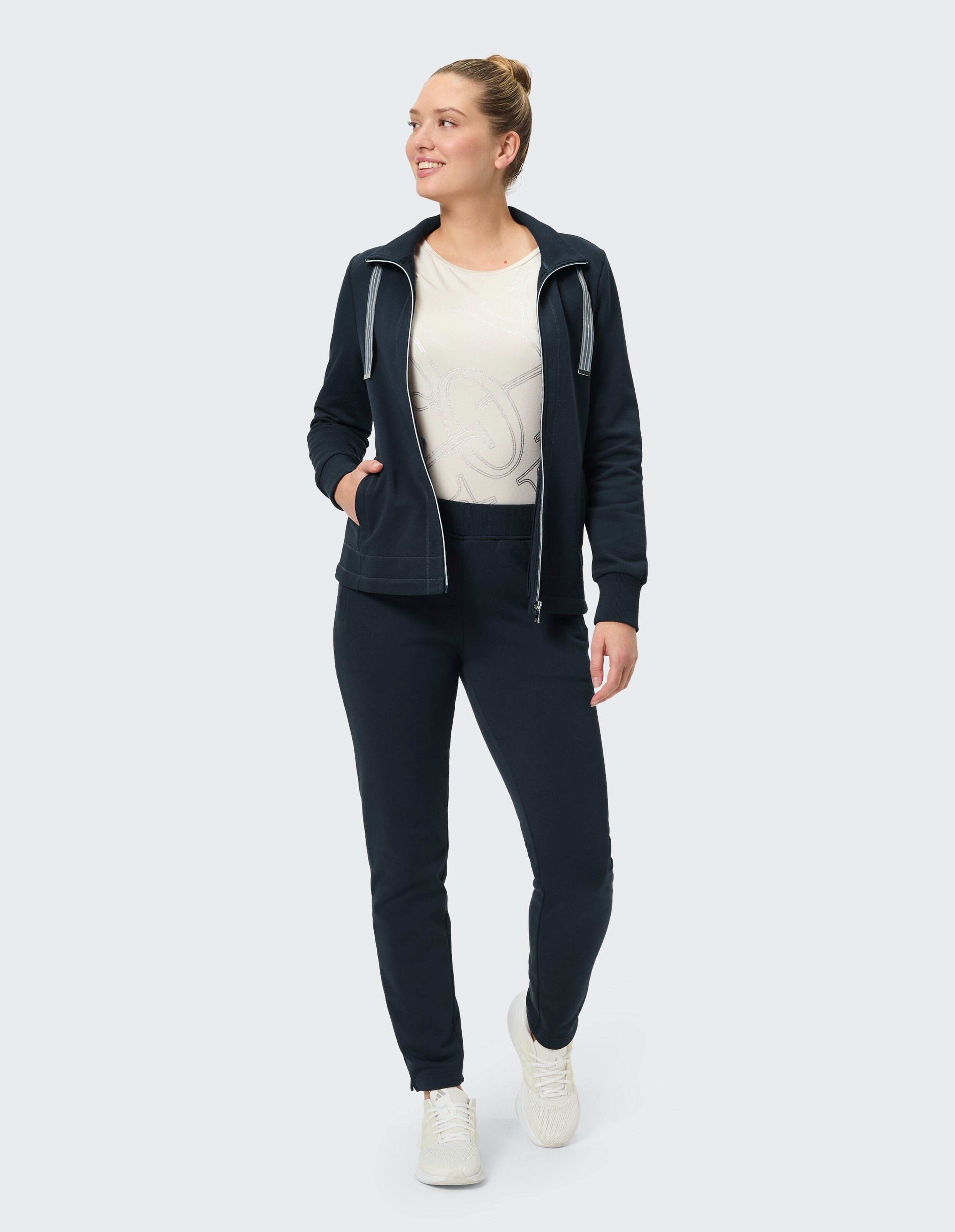 Frau in dunkelblauem Trainingsanzug und rosa T-Shirt mit Aufdruck, steht in lässiger Pose, schaut nach links gerichtet.