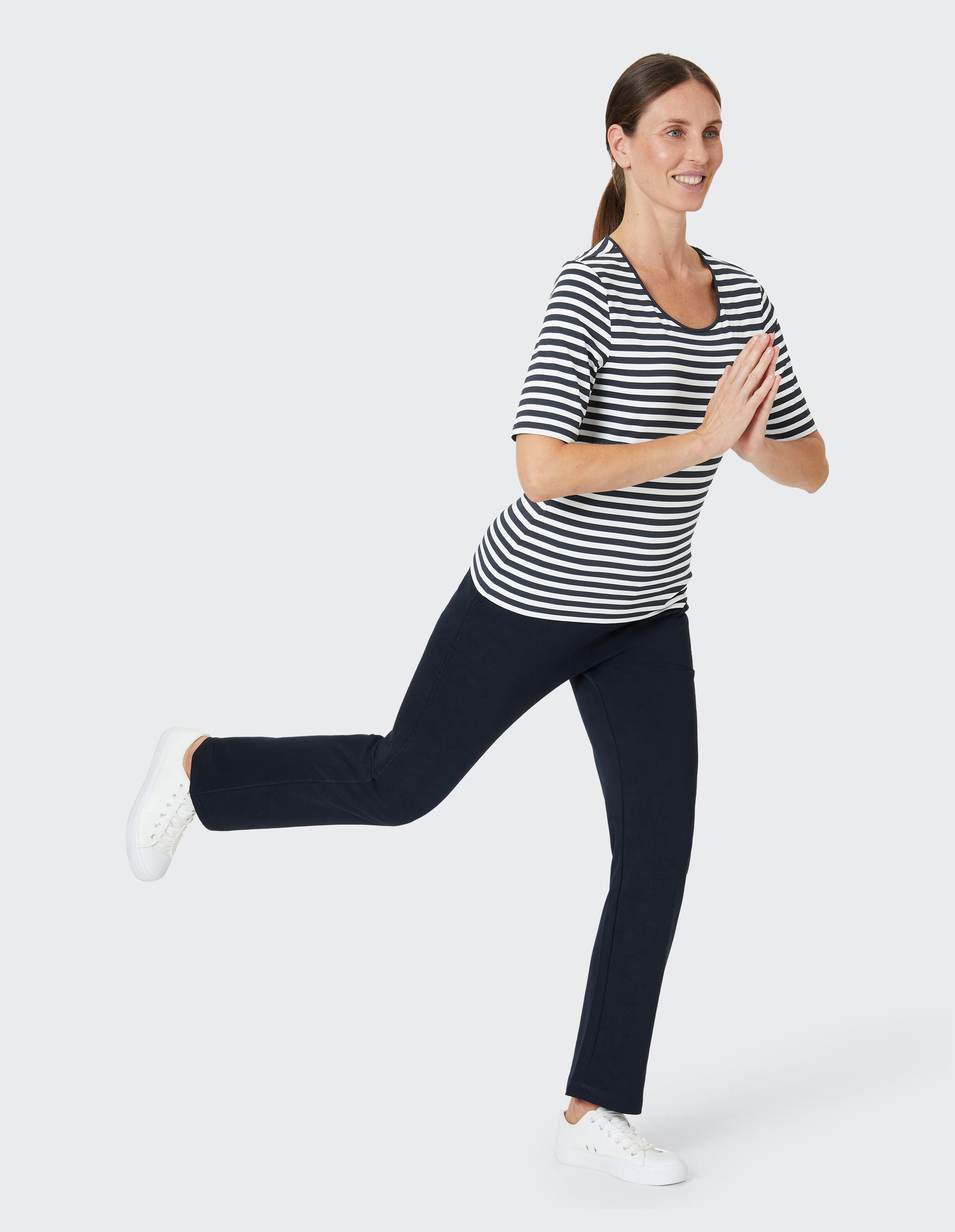 Frau in gestreiftem Shirt und dunkler Hose balanciert auf einem Bein, Hände betend gefaltet, lächelnd, vor hellem Hintergrund.