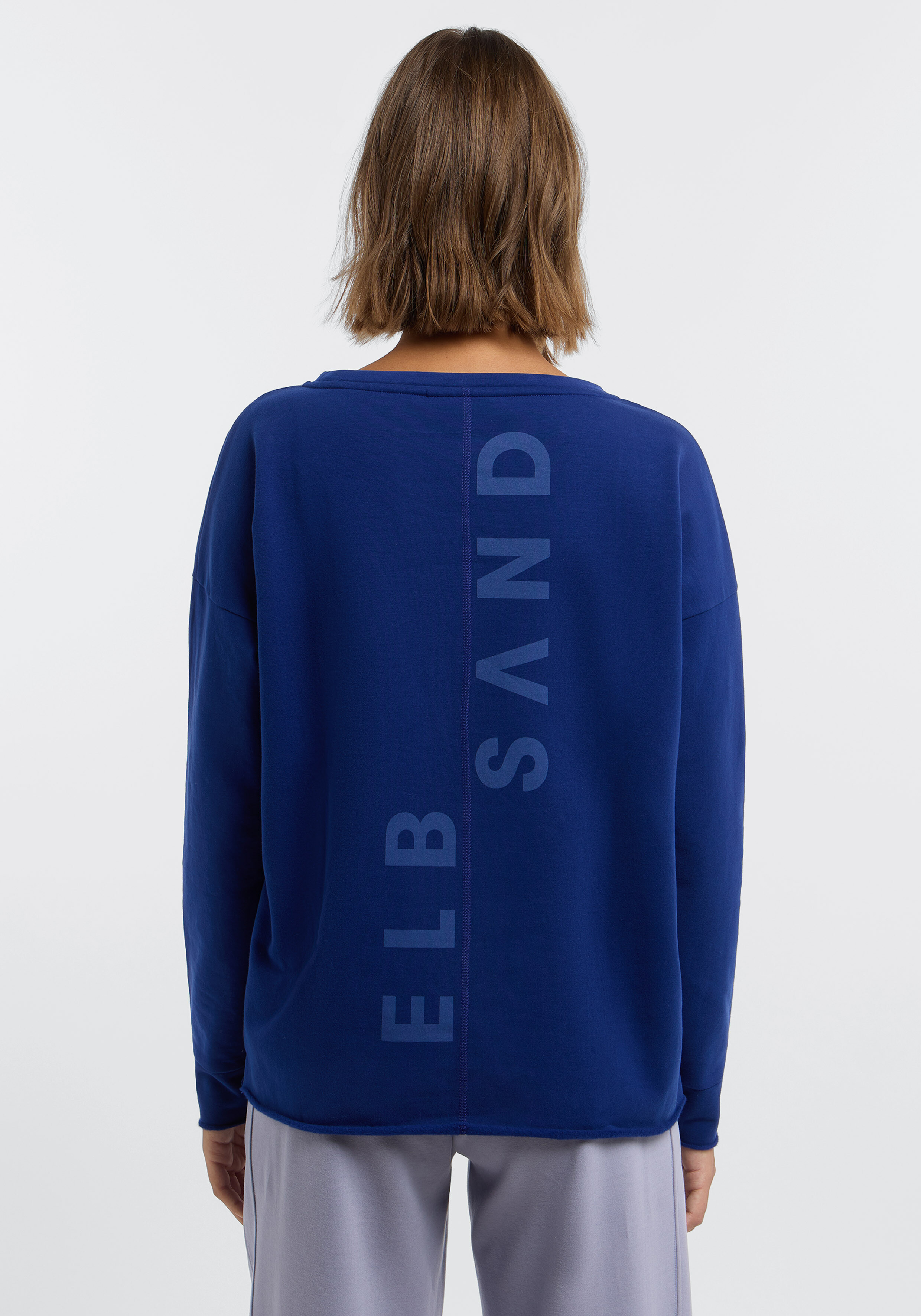 Frau trägt ein kuscheliges blaues ELBSAND-Sweatshirt mit Logo-Aufdruck auf der Rückseite für lässige Loungewear.