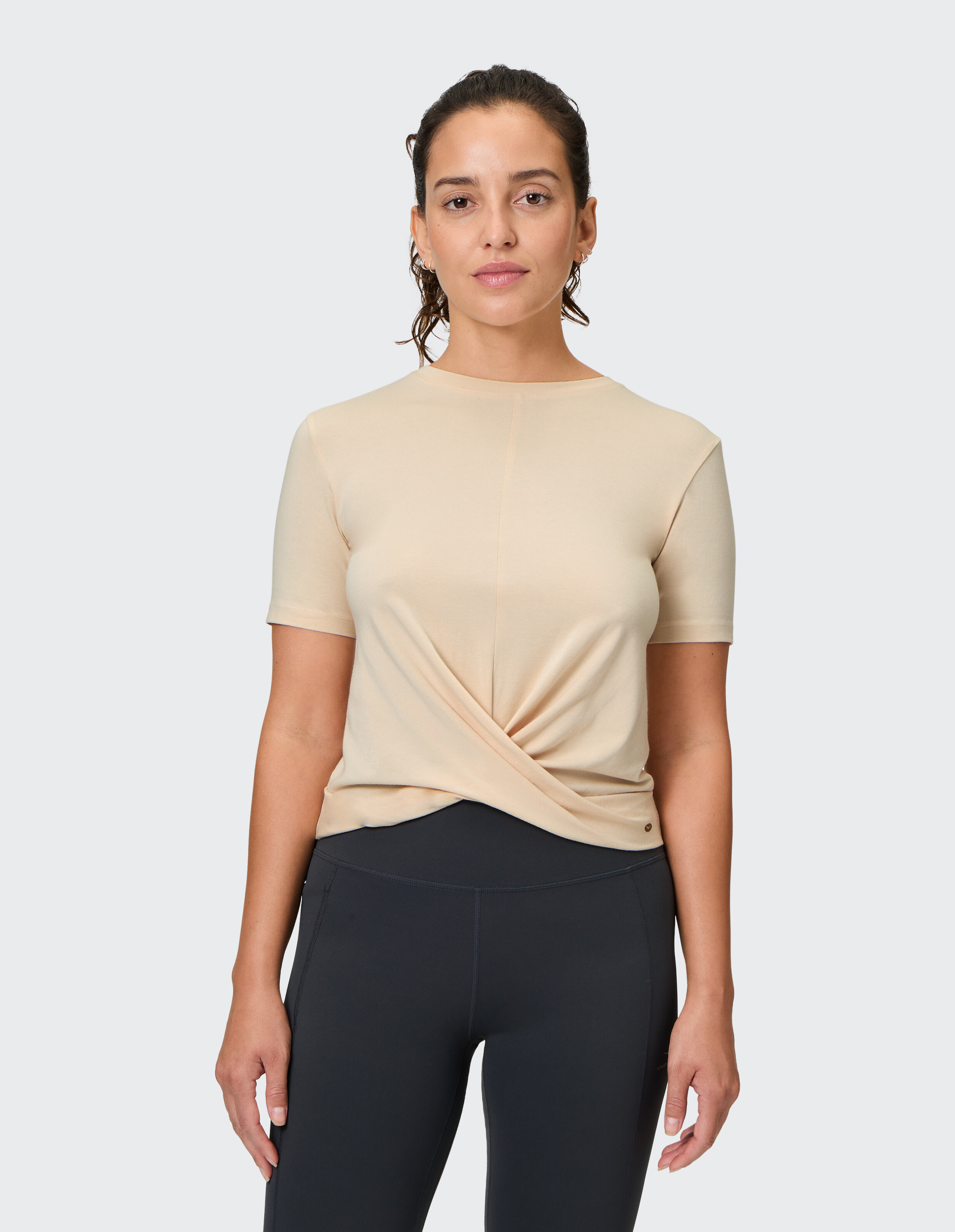 Frau in einem beigen T-Shirt mit verdrehter Vorderseite gepaart mit schwarzen Leggings.