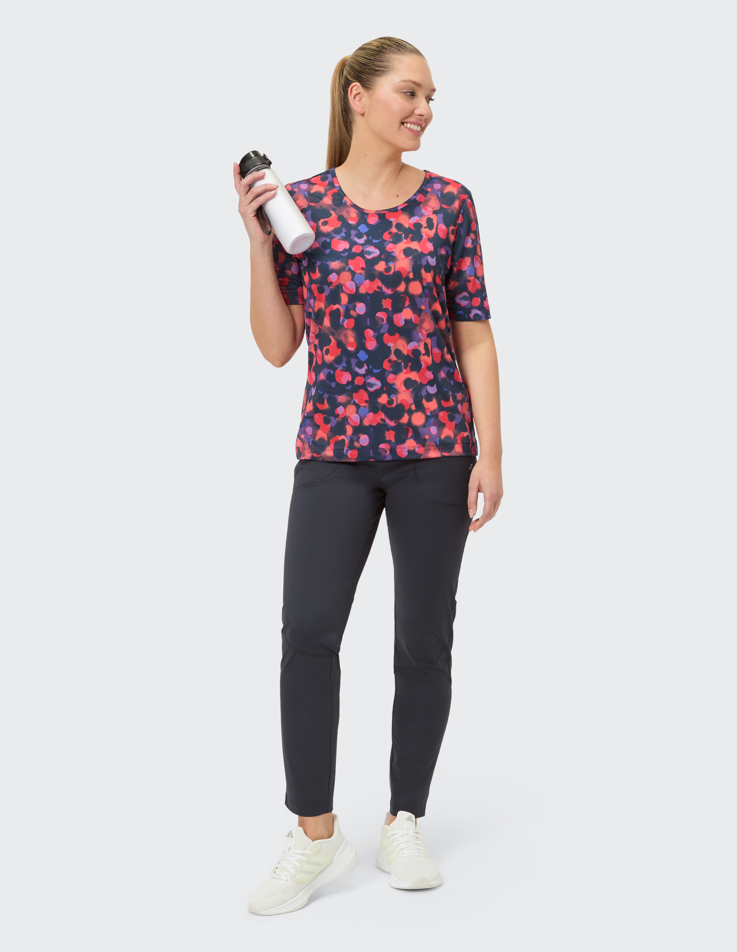 Frau in bunt gemustertem Oberteil und dunkler Hose, hält eine Wasserflasche in der Hand.