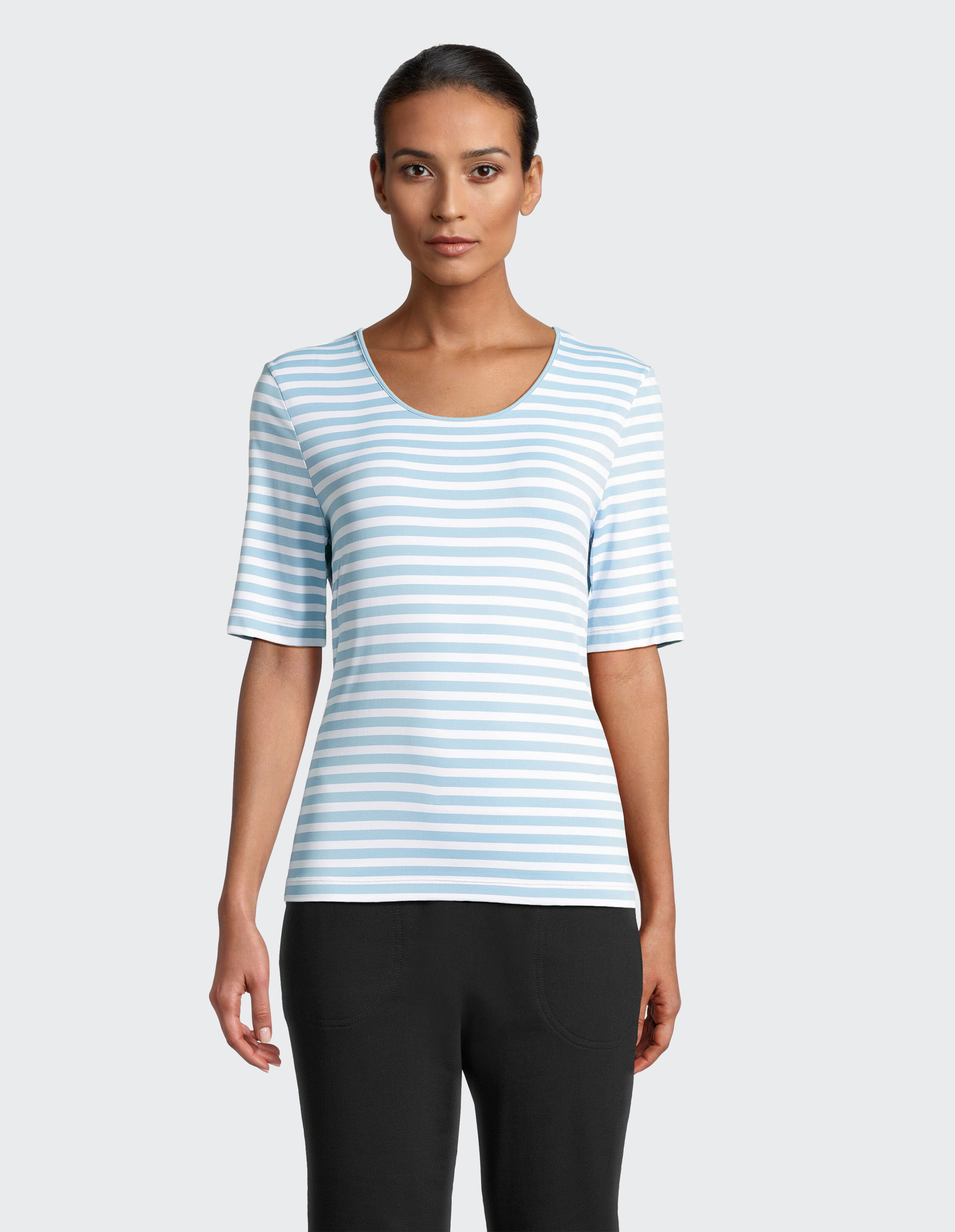 Frau in hellblau-weiß gestreiftem T-Shirt mit kurzen Ärmeln und schwarzen Hosen vor grauem Hintergrund.
