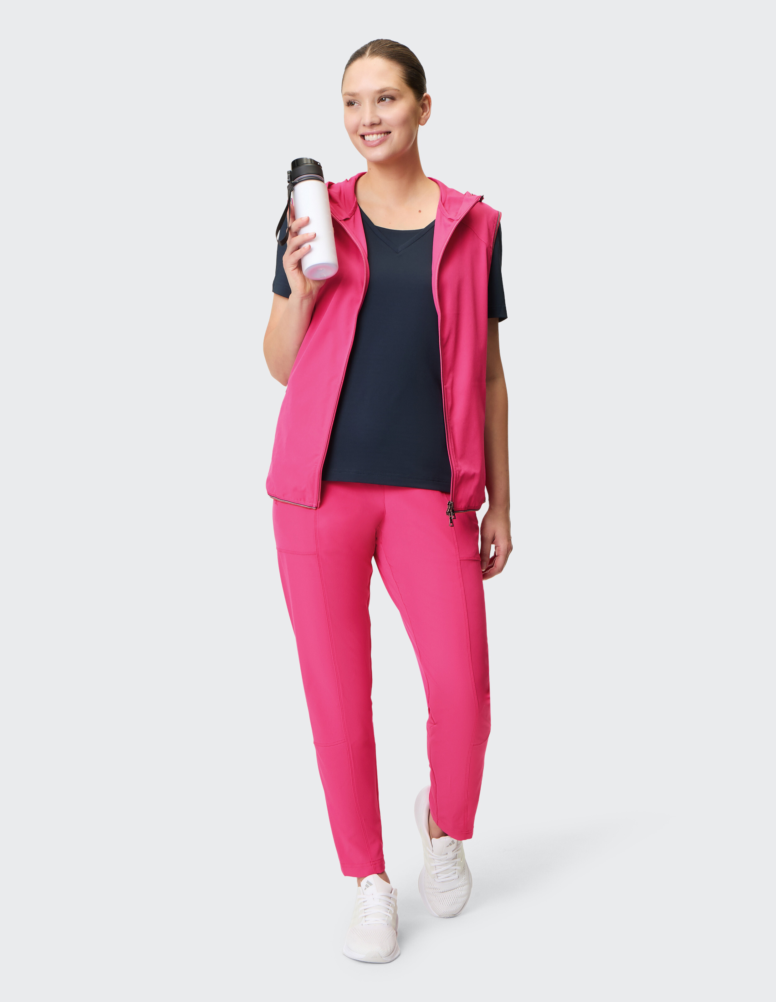 Frau in leuchtend rosa Sportoutfit mit Kapuzenweste und passender Hose, hält eine Wasserflasche in der Hand und lächelt.