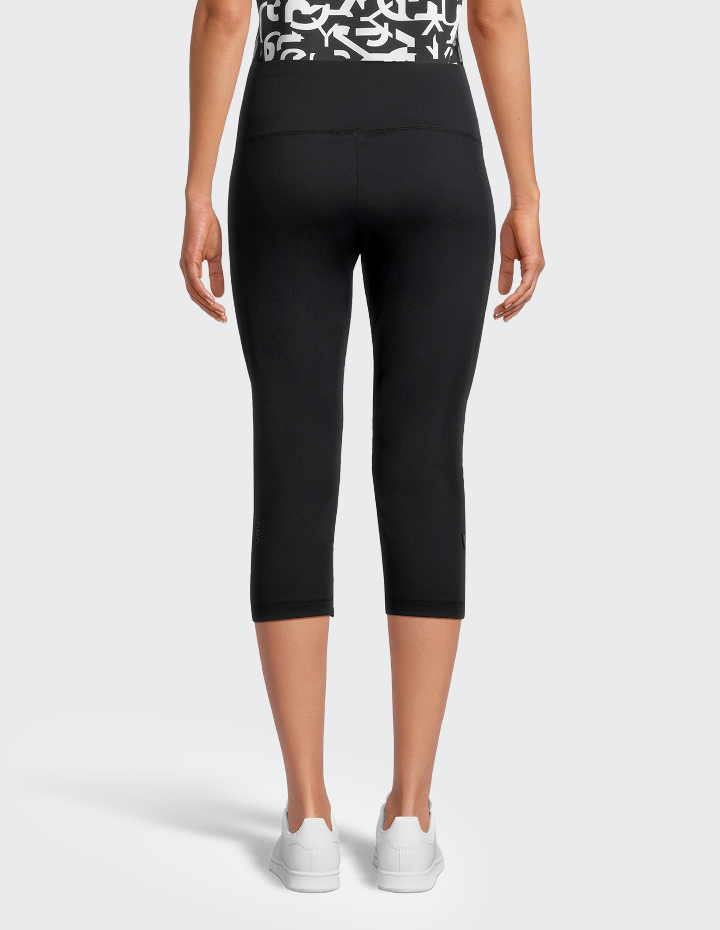 Der Rückenansicht einer Person in schwarzer knielanger Leggings, weißem Muster-Oberteil und weißen Sneakern auf grauem Hintergrund.