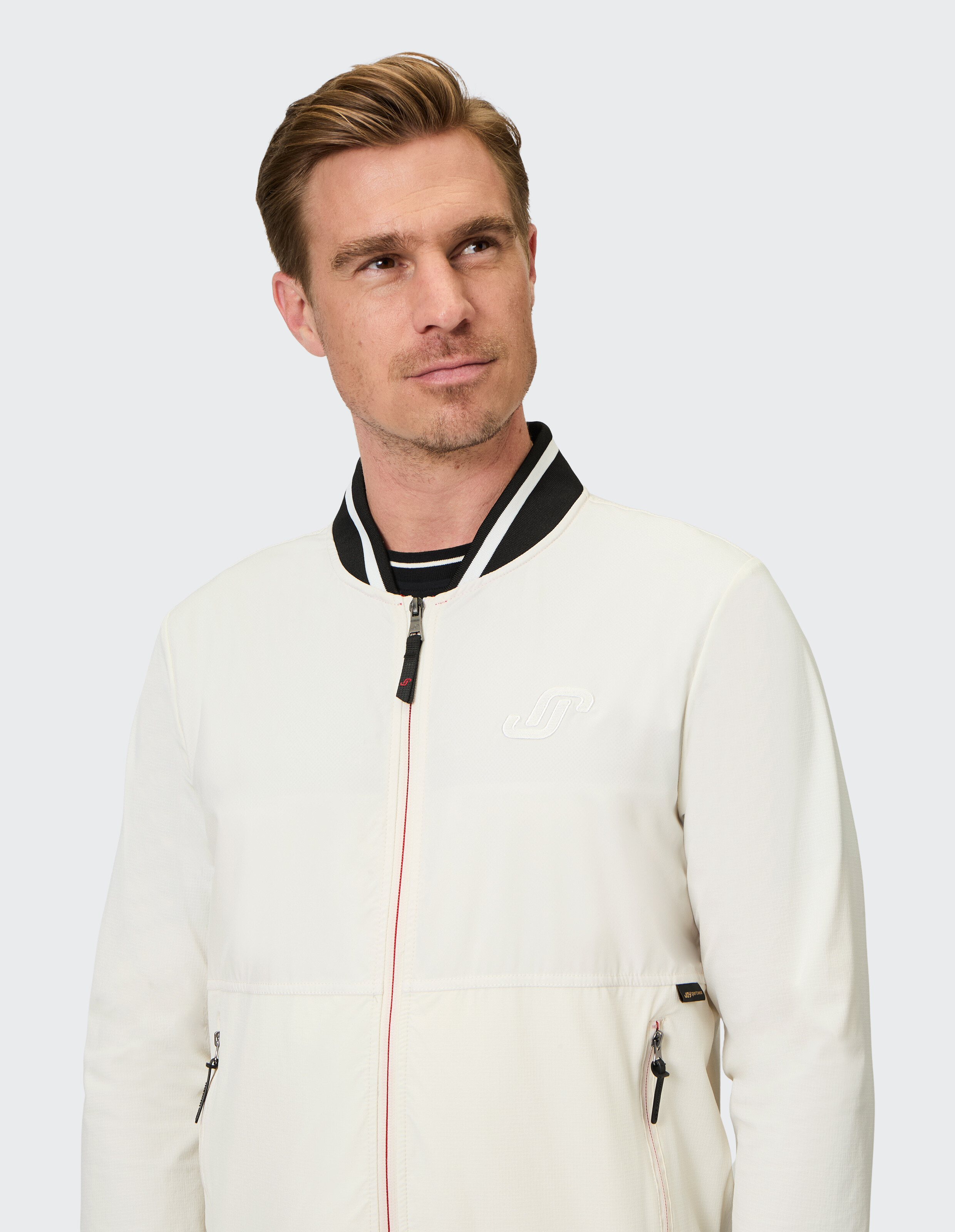 Mann mit blondem Haar trägt eine weiße Jacke mit schwarzem Kragen und Logo und blickt nach rechts vor neutralem Hintergrund.