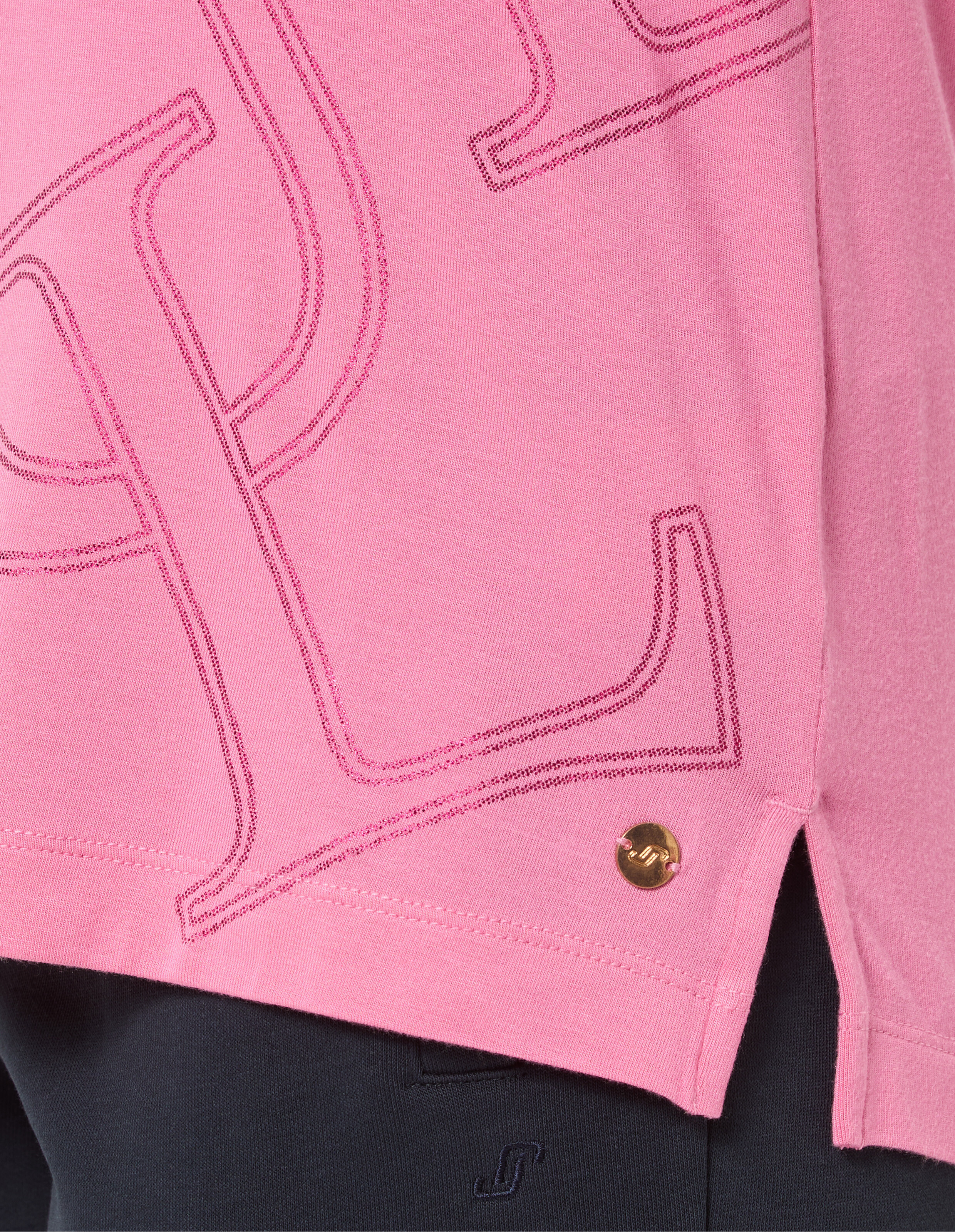 Rosa Sweatshirt mit dunkelrosa gesticktem Muster und goldener Metallniete am Saum, kombiniert mit schwarzer Hose darunter.