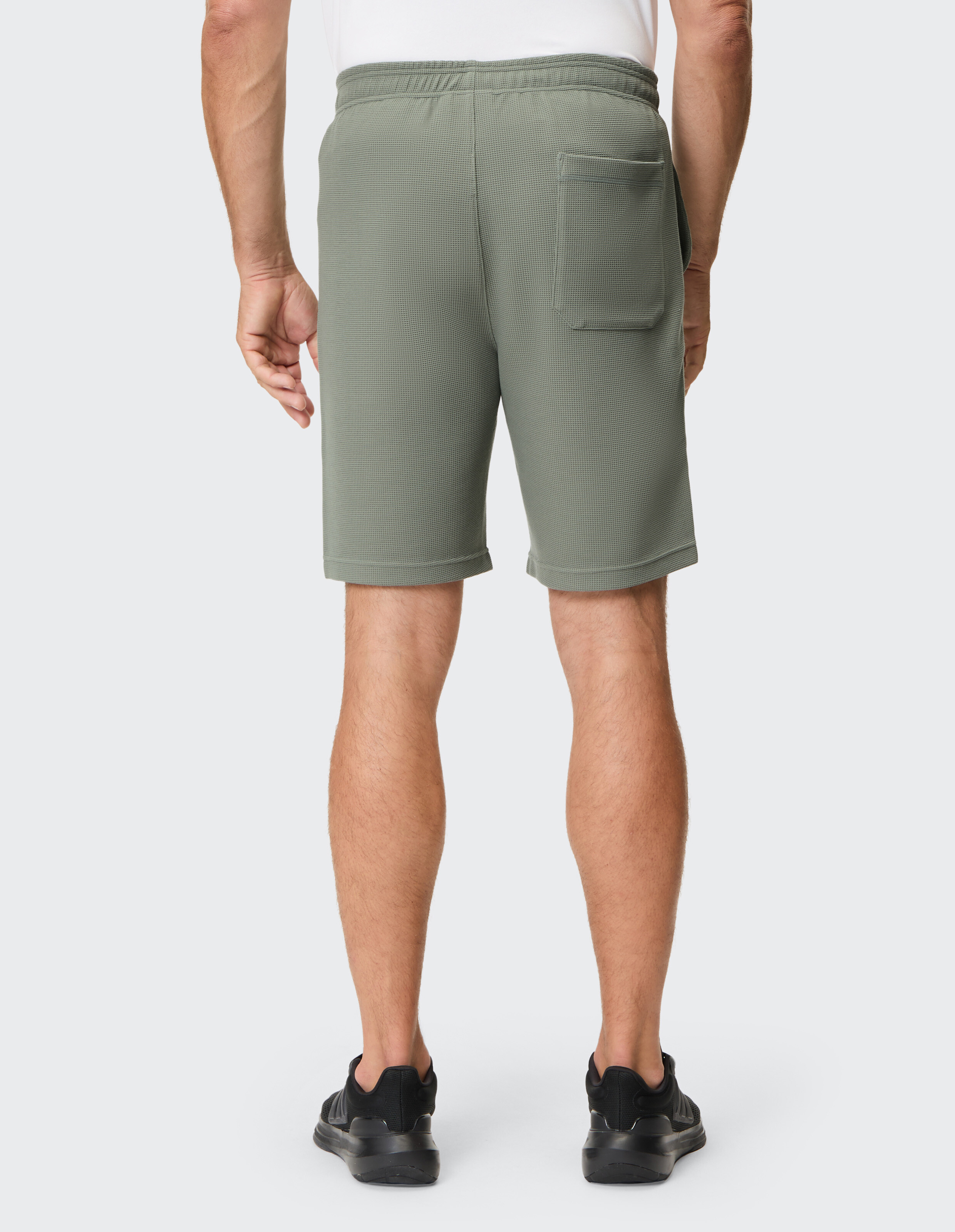 Ein Mann in olivgrünen Shorts mit elastischem Bund und Rückentasche, schwarzem Schuhwerk, steht vor einem weißen Hintergrund.