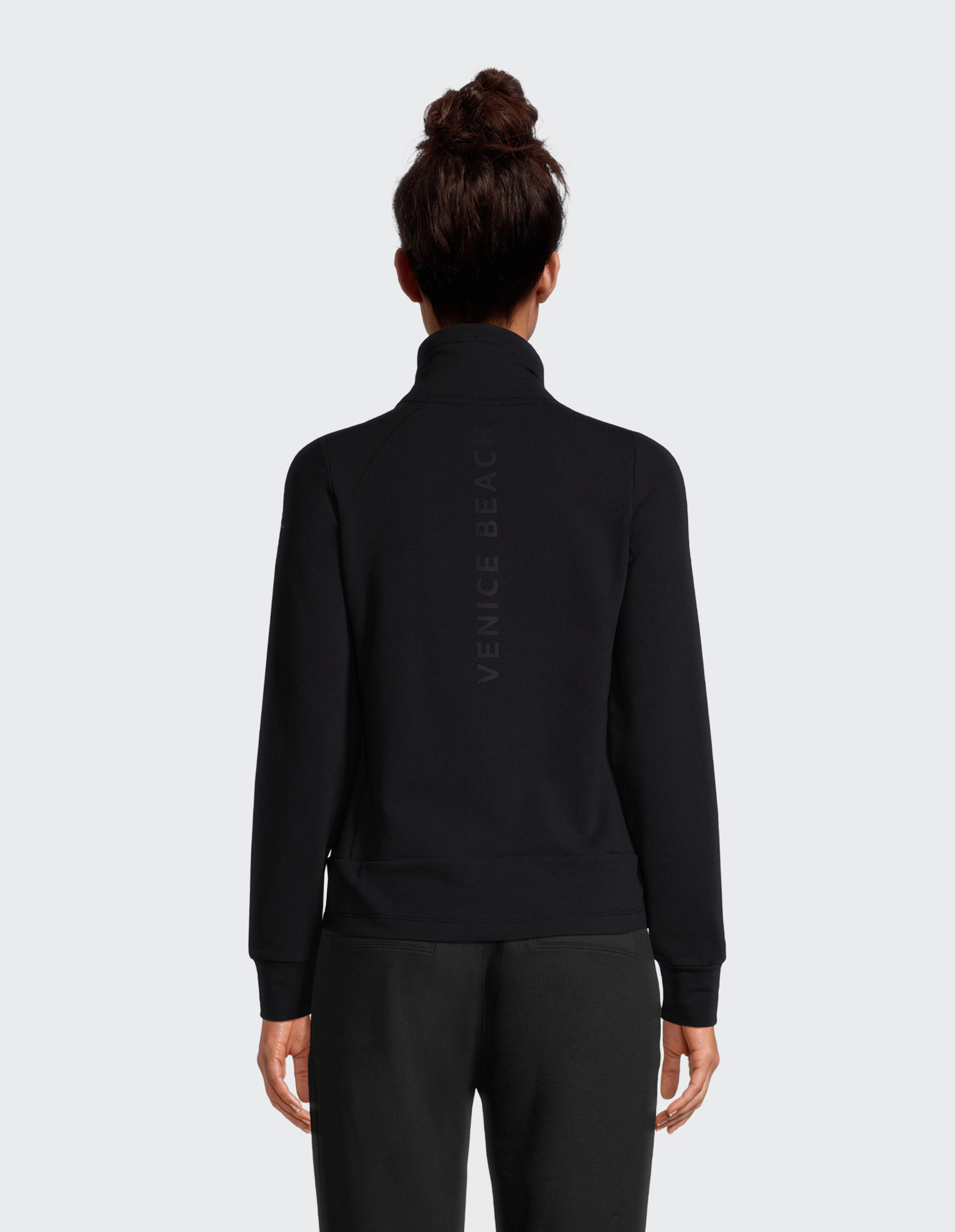 Rückansicht einer Frau, die eine schwarze Freizeitjacke mit Reißverschluss und Venice Beach-Branding trägt.