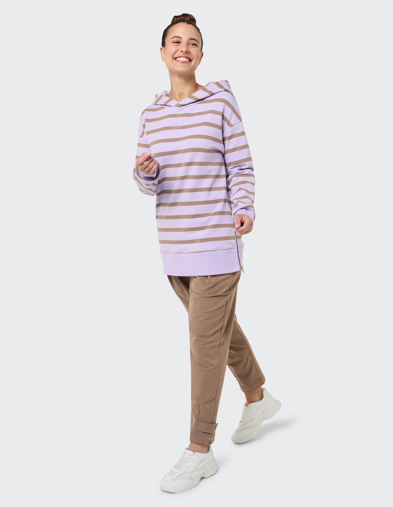Lächelnde Frau in einem gestreiften lavendelfarbenen Kapuzenpullover und einer braunen Hose, gestylt für Freizeitkleidung.