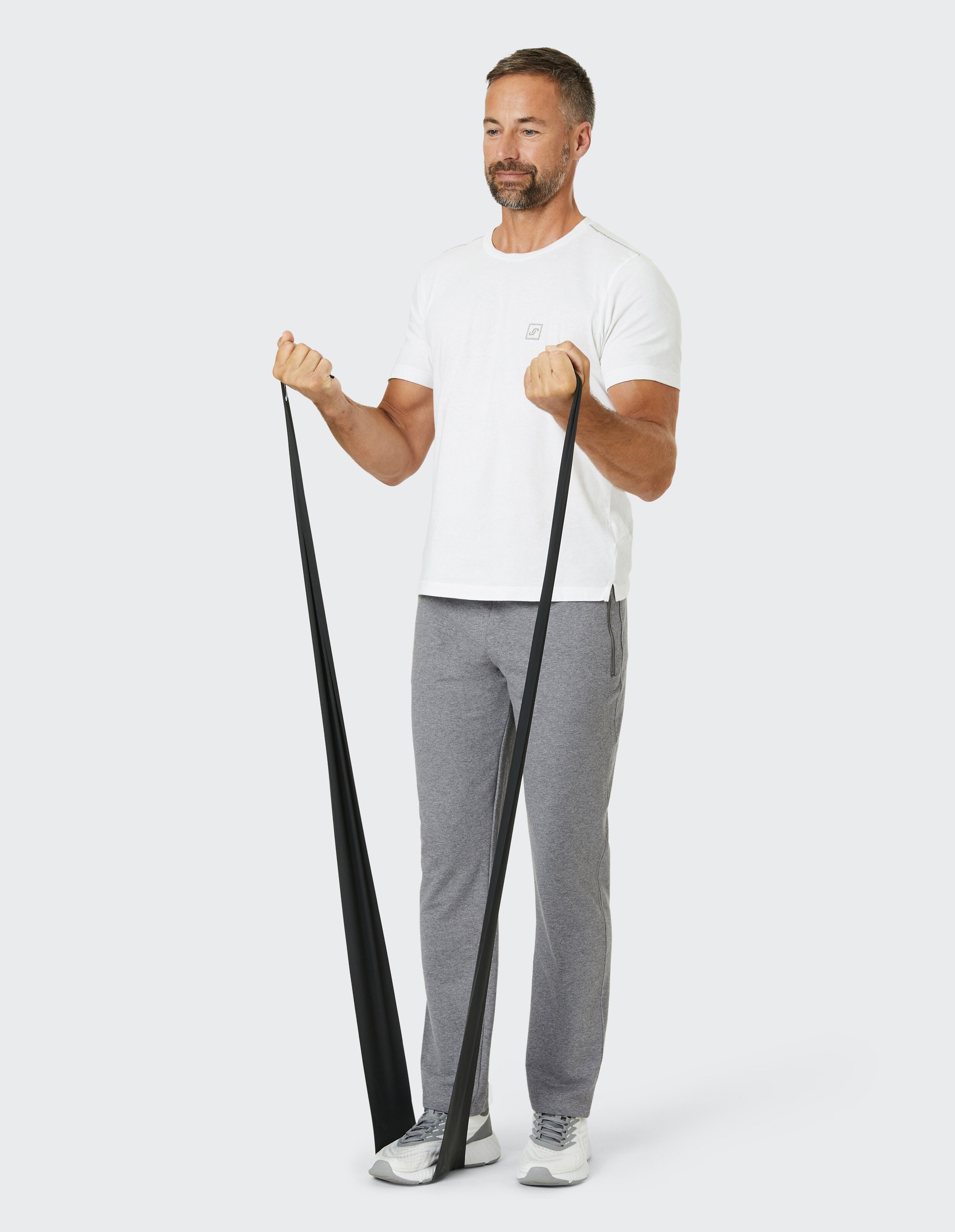 Ein Mann in weißem T-Shirt und grauer Jogginghose trainiert mit einem schwarzen Widerstandsband vor einem neutralen Hintergrund.