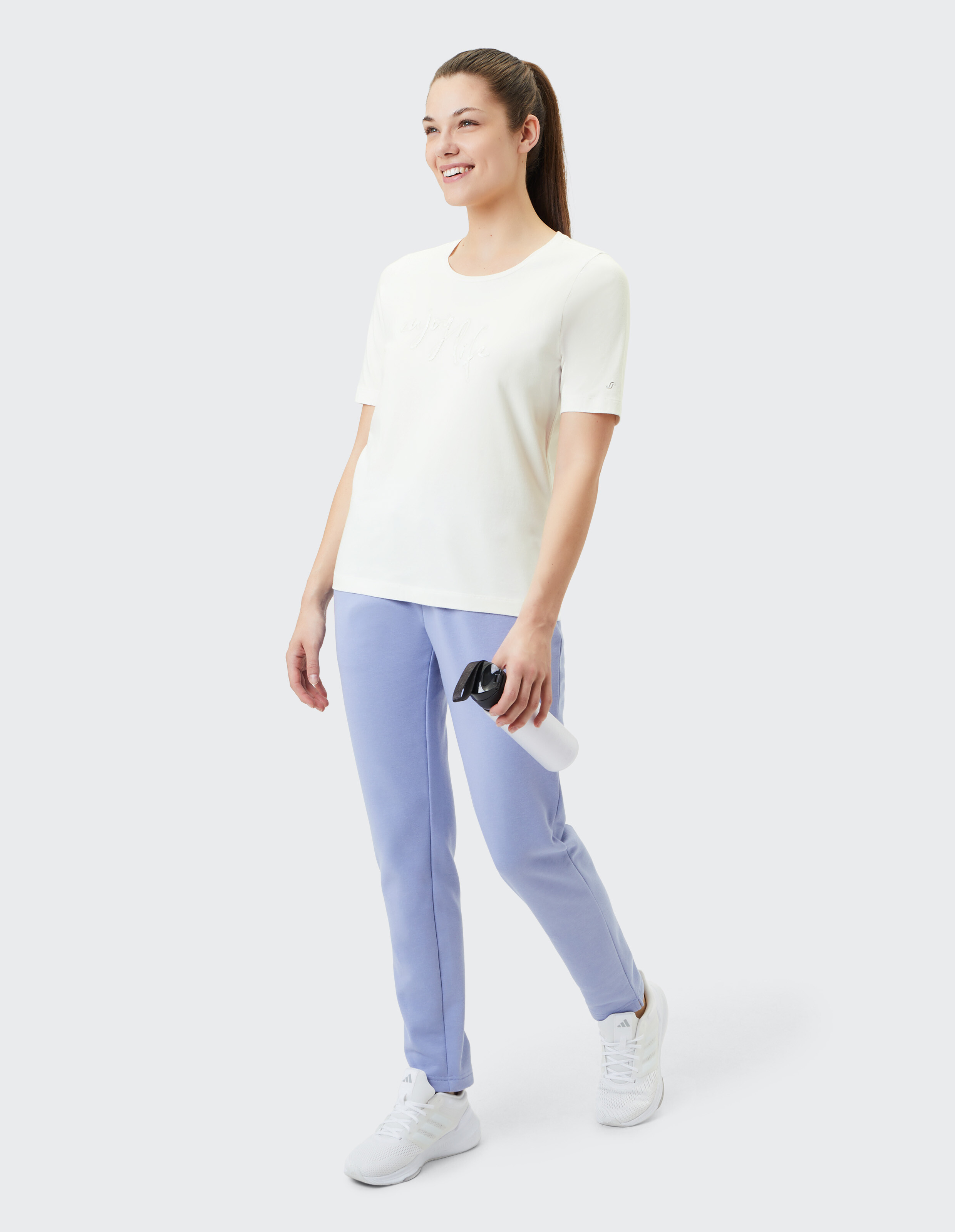 Frau in weißem T-Shirt und lavendelfarbener Jogginghose hält eine Wasserflasche in der Hand.