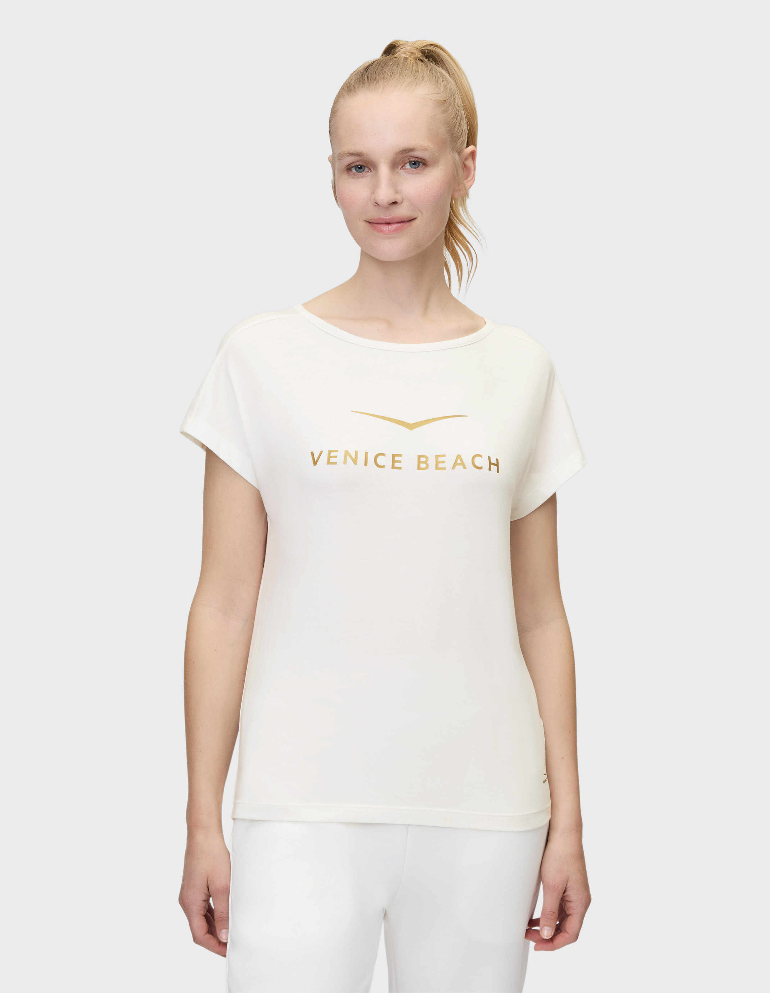 Frau trägt ein weißes, lässiges T-Shirt mit goldenem Venice Beach-Logoaufdruck.