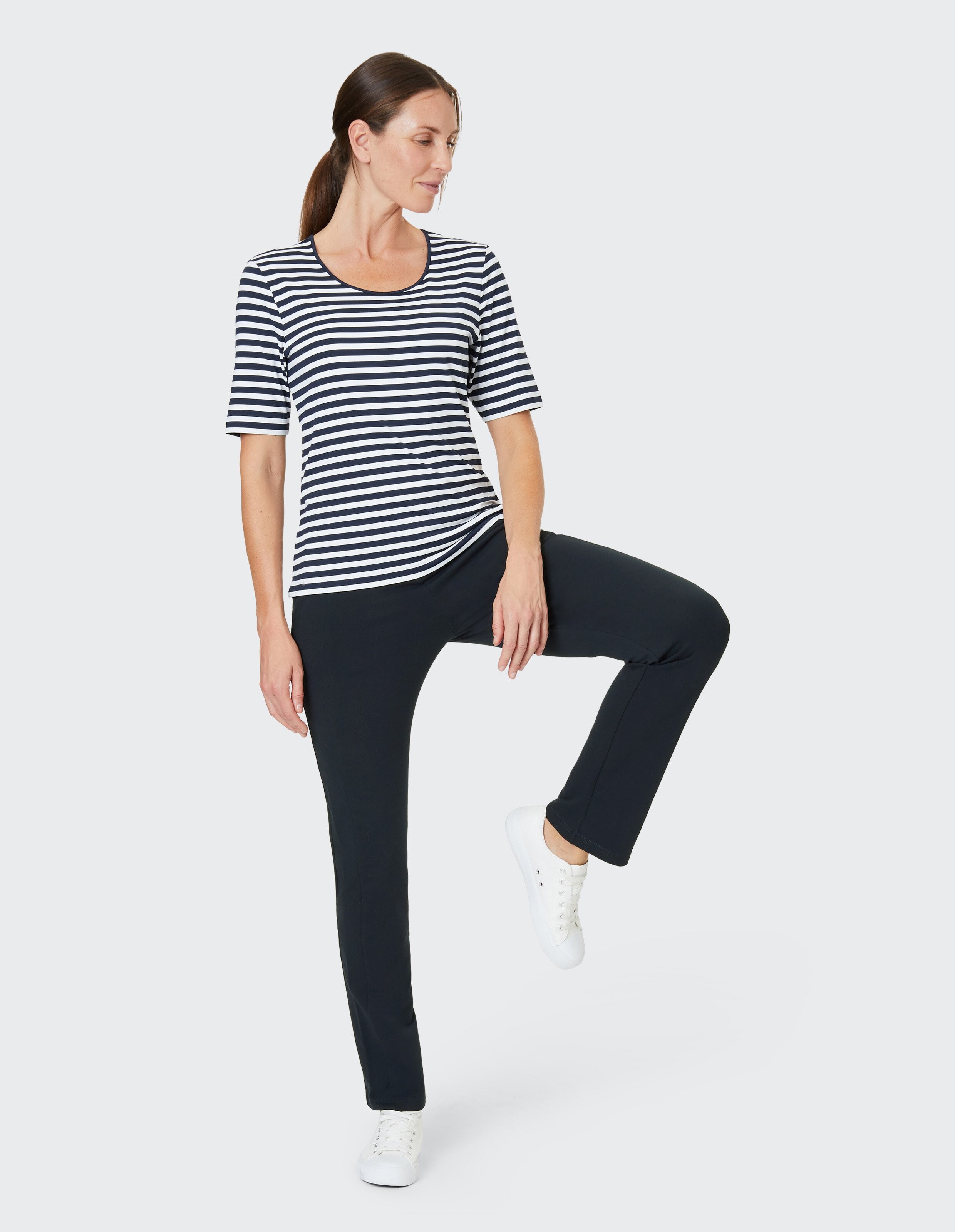 Frau in gestreiftem Shirt und schwarzer Hose hebt ein Bein, trägt weiße Sneakers, vor weißem Hintergrund.