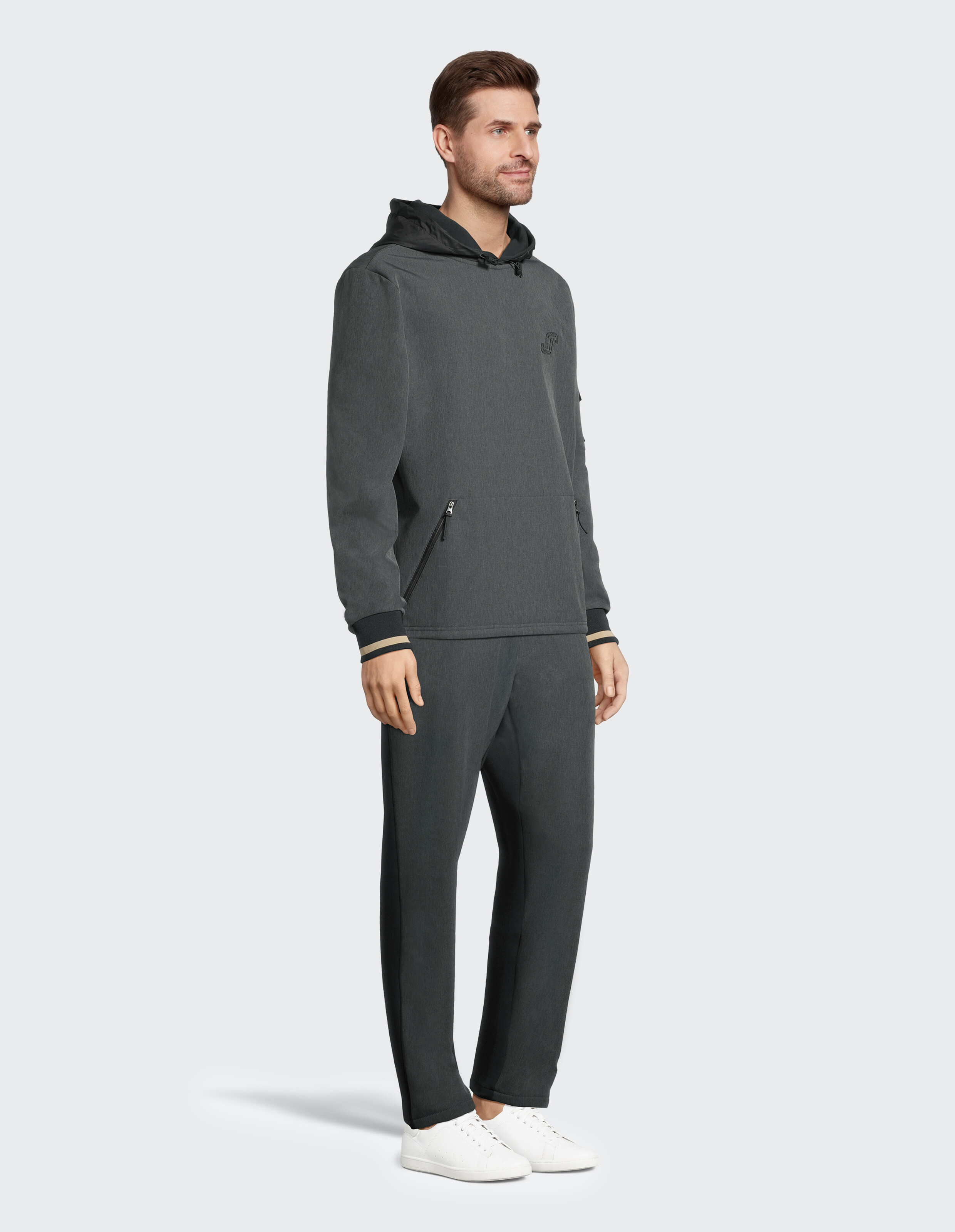 Ein Mann trägt einen dunkelgrauen Hoodie und Jogginghose mit weißen Sneakers vor weißem Hintergrund, lächelnd in Dreiviertelansicht.