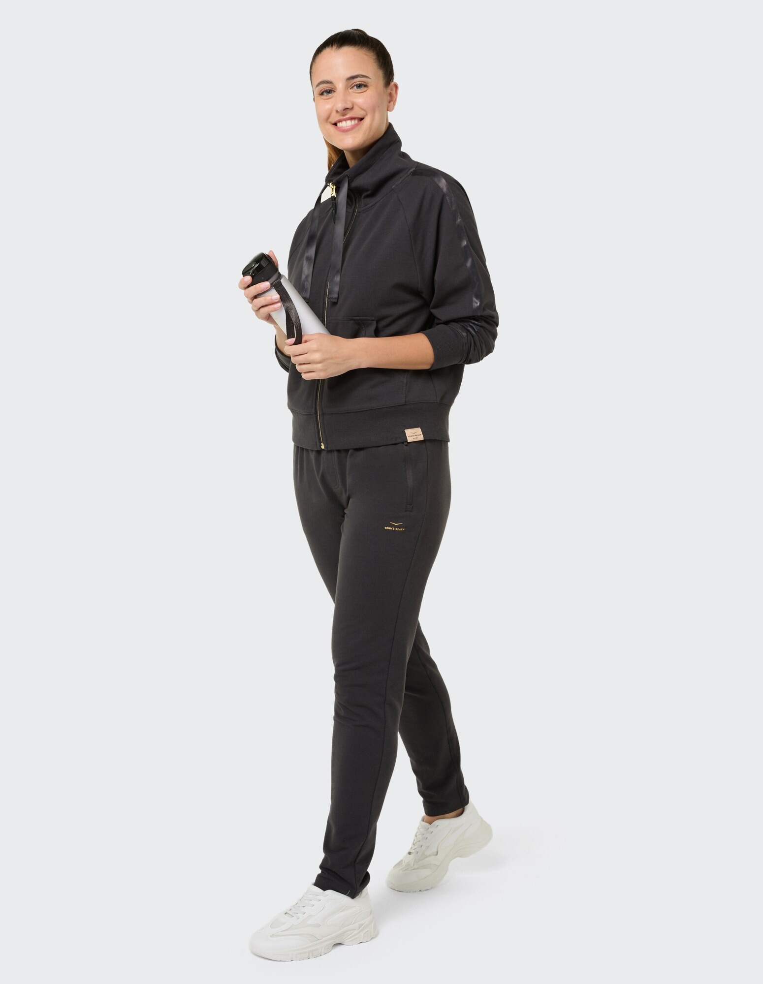 Frau in einem stylischen schwarzen Trainingsanzug, hält eine Wasserflasche in der Hand und lächelt.