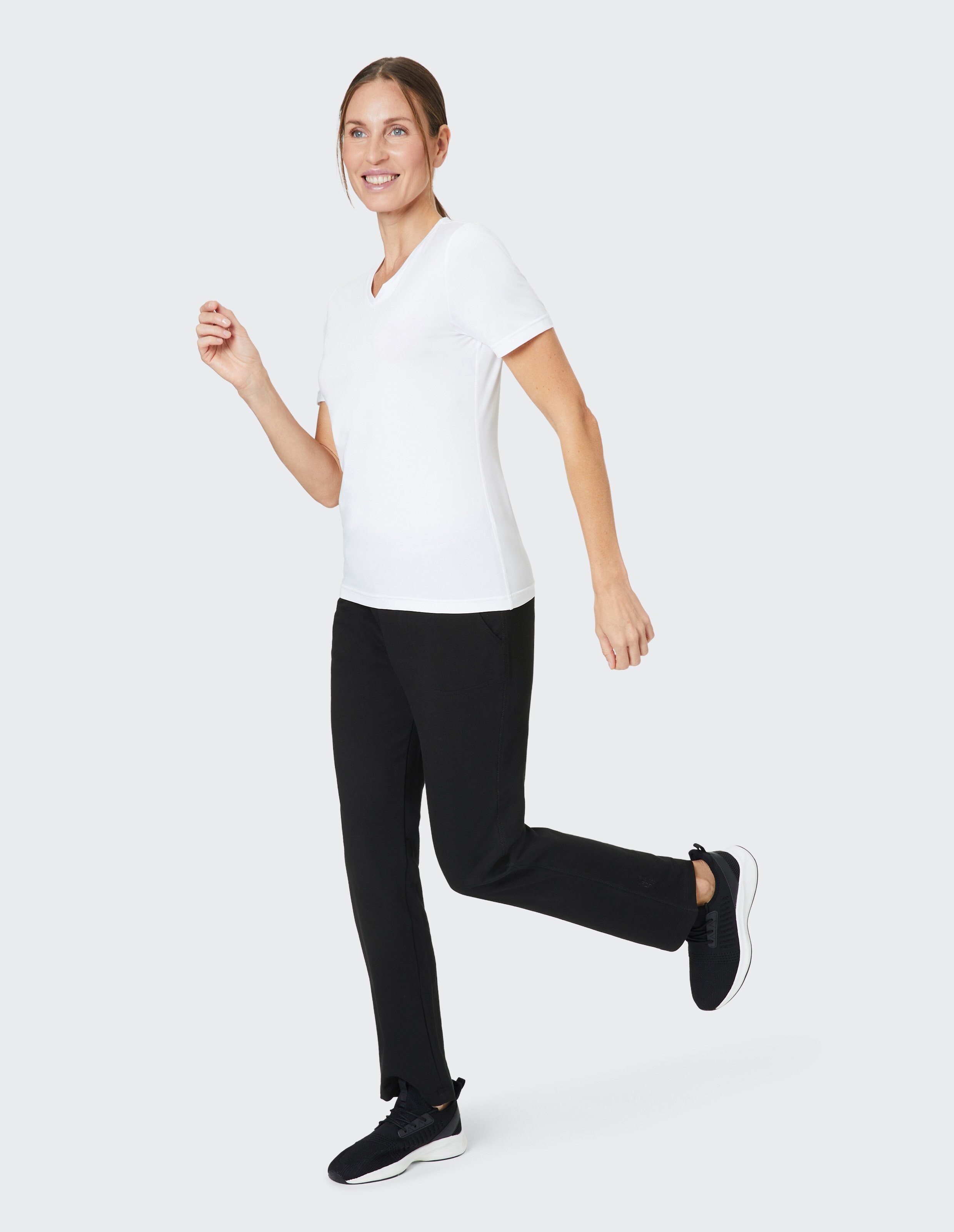 Frau in weißem T-Shirt, schwarzer Hose und Sneakern auf grauem Hintergrund, lächelnd und in einer gehenden Pose.