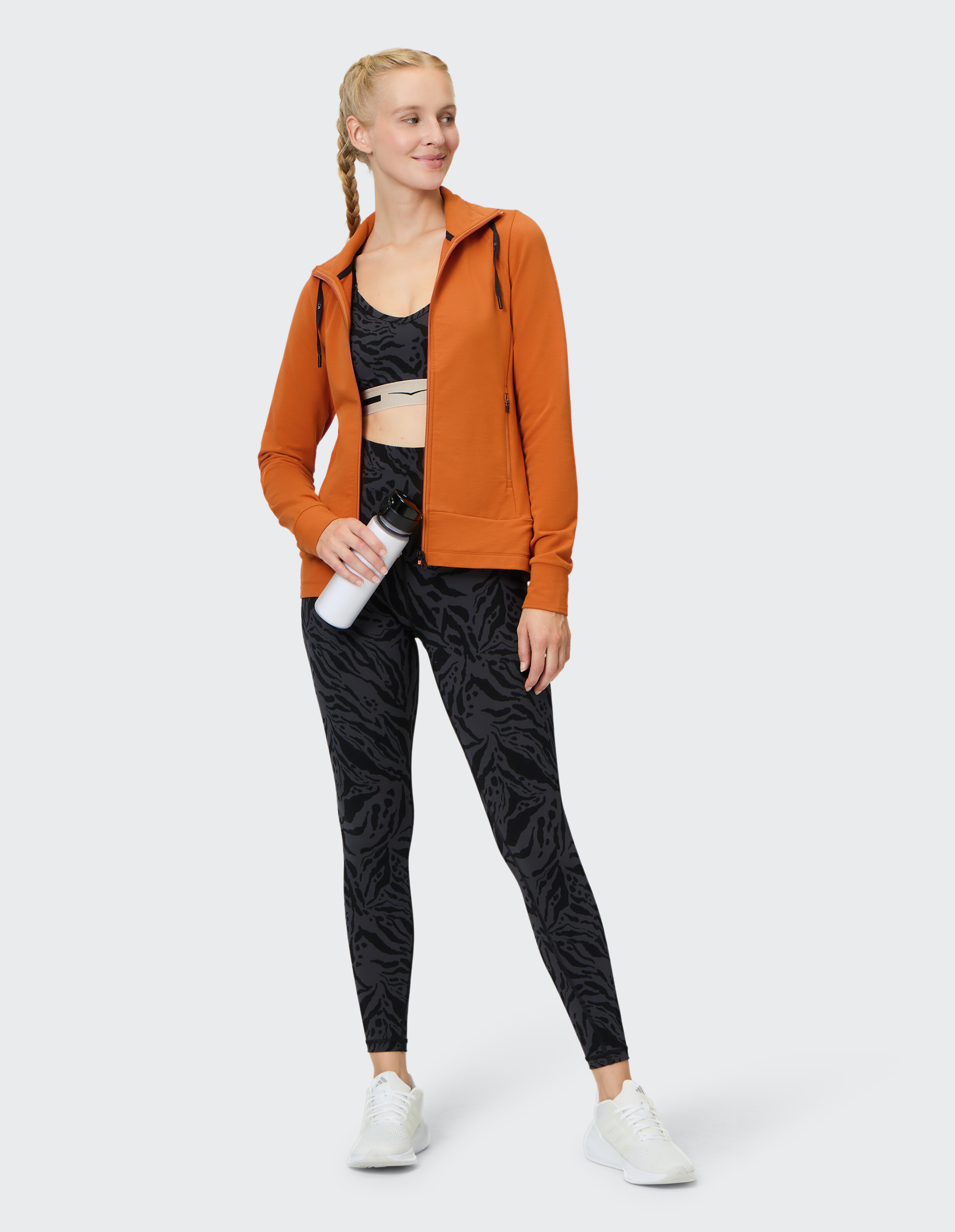 Frau in orangefarbenem Kapuzenpullover mit Reißverschluss, schwarzen Leggings mit Animal-Print und Sport-BH für aktive Freizeitkleidung.