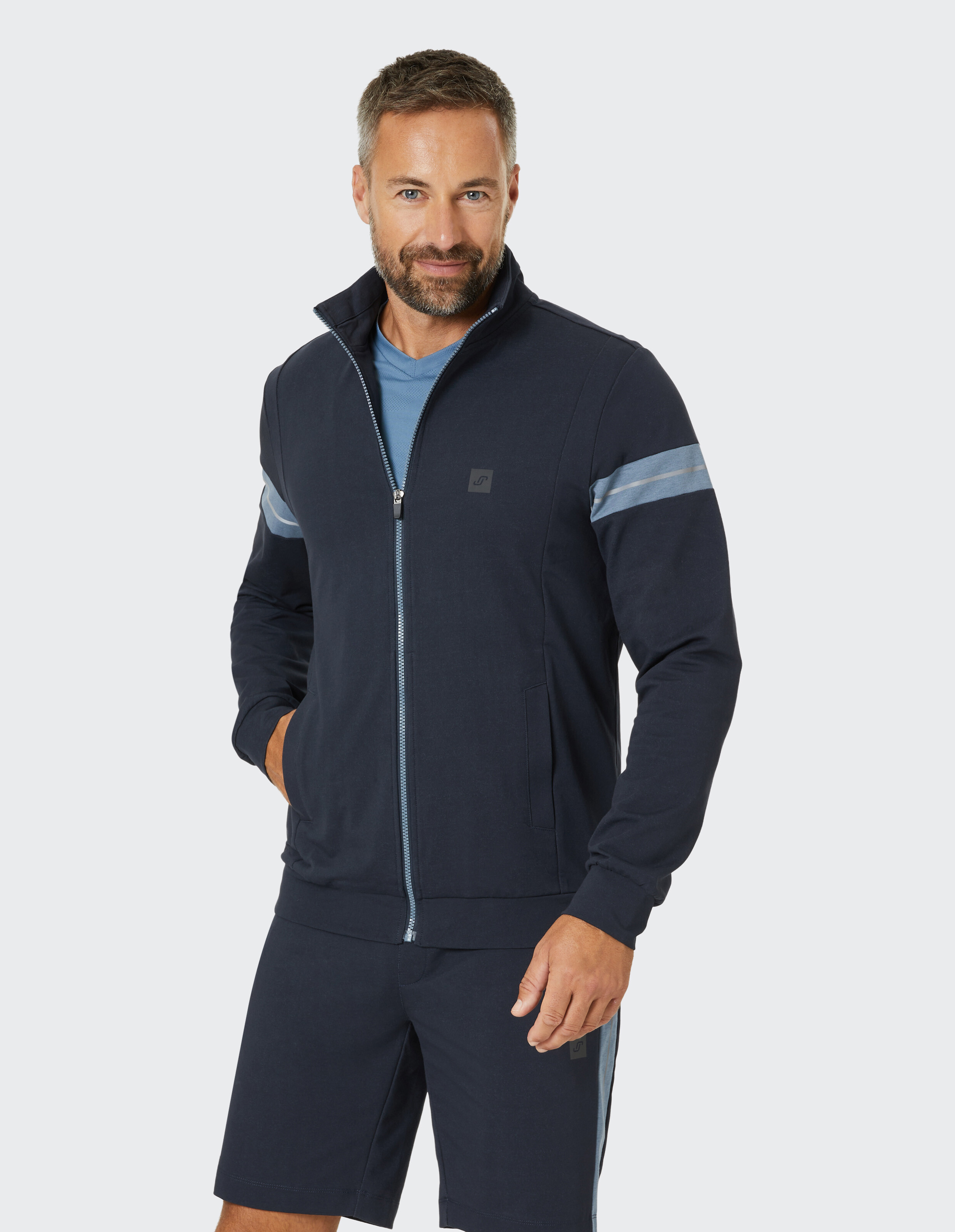 Ein Mann trägt eine dunkelblaue Sportjacke mit Reißverschluss und passender Shorts, stehend vor einem hellen Hintergrund.